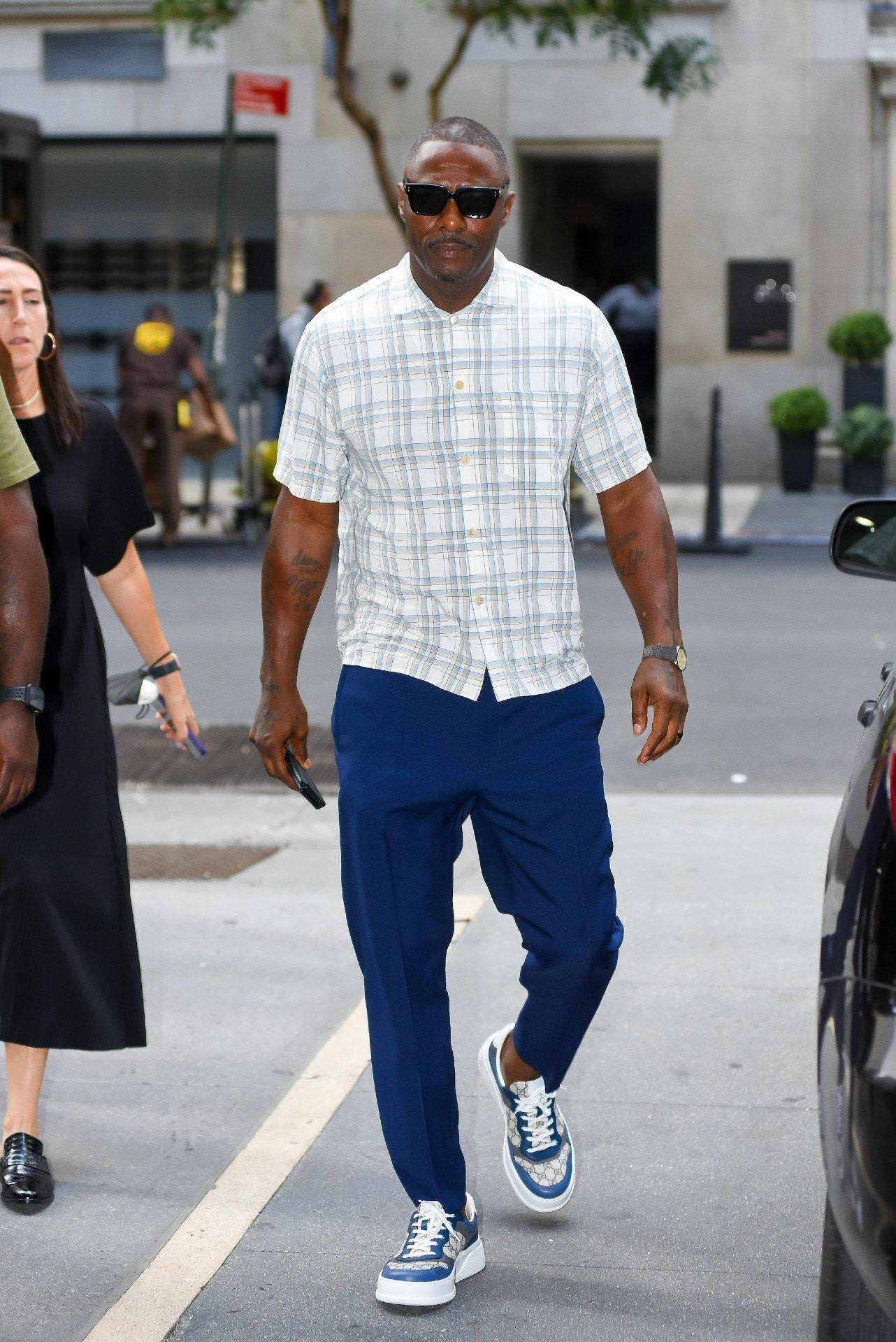 Celebrity 桌布 壁紙 — 一位時尚男士穿著格紋襯衫和海軍藍長褲，戴著太陽眼鏡和圖案運動鞋，自信地走在城市街道上。