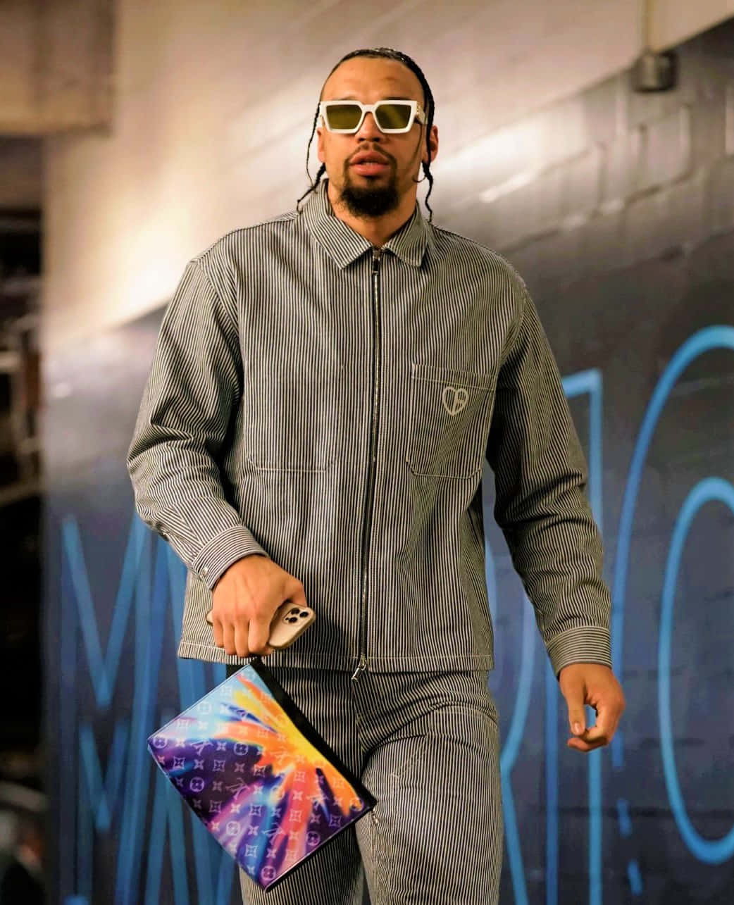Dillon Brooks 桌布 壁紙 — 一位時尚男士穿著條紋工作服和白色太陽眼鏡，自信地走著，手持色彩繽紛的手拿包和手機。