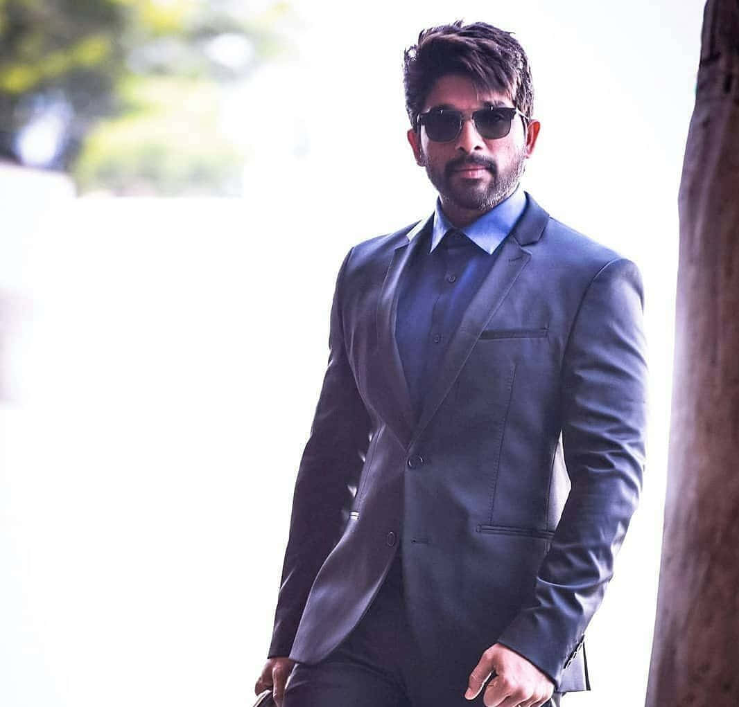 Allu Arjun 桌布 壁紙 — 一位時尚男士穿著深色西裝和太陽眼鏡，自信地站在明亮的戶外背景前，背景有模糊的綠色植物。