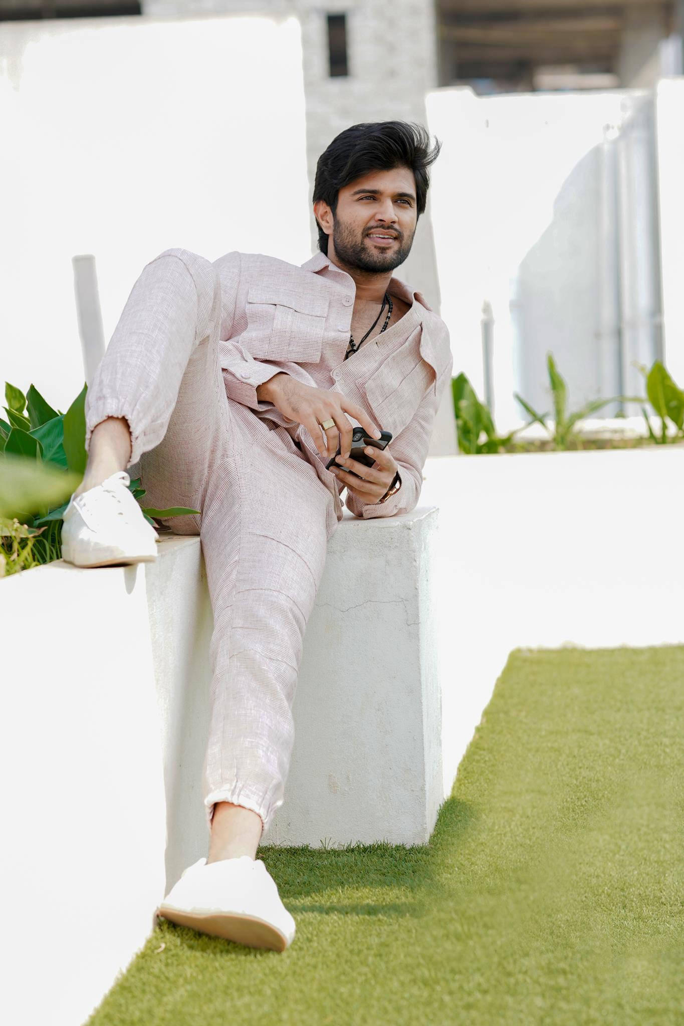 vijay devarakonda 4K 桌布 壁紙 — 一位時尚男士穿著淺粉紅色亞麻布，坐在白色混凝土邊緣上，手持電話，背後有綠色草地和現代建築。