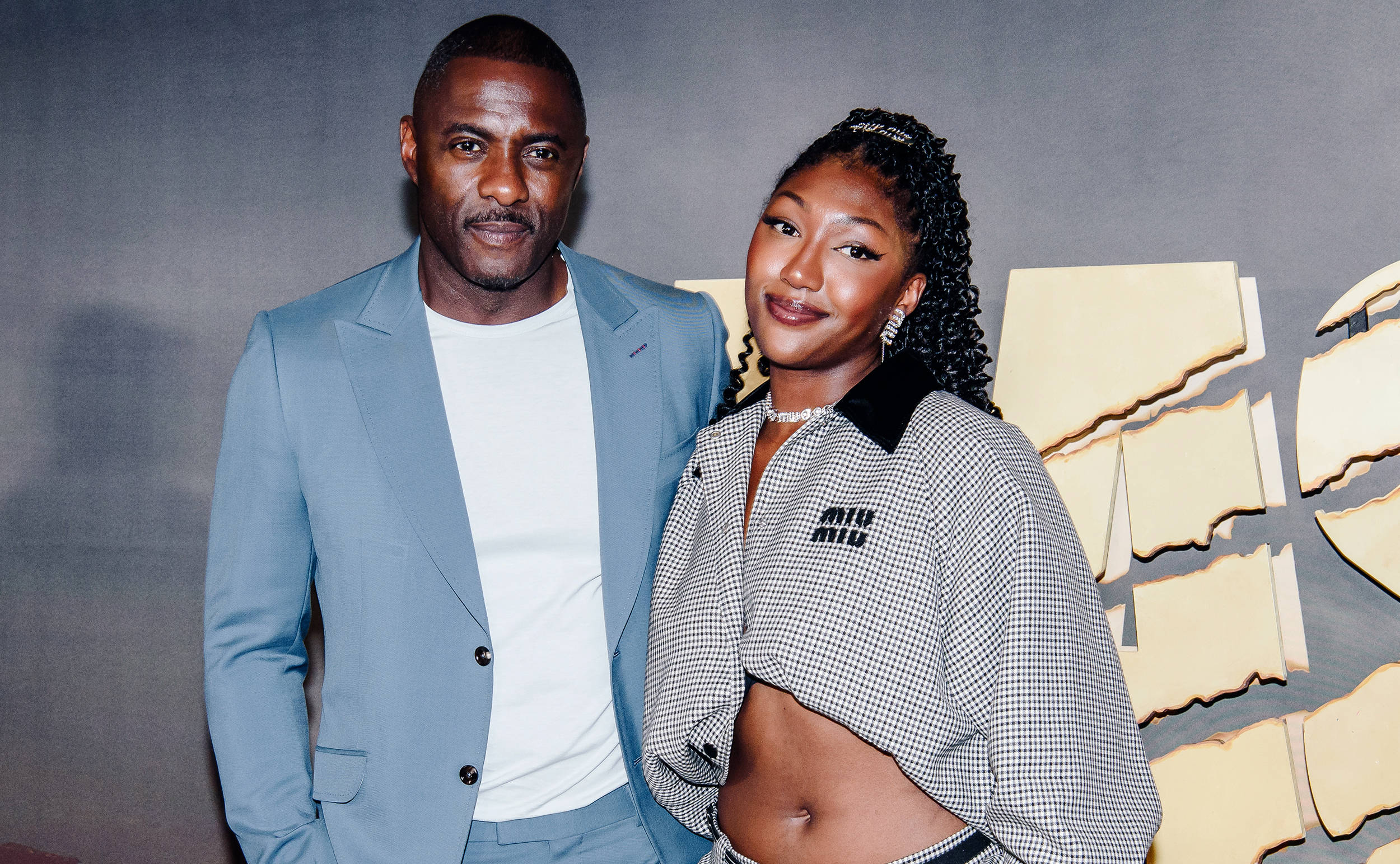 idris elba 桌布 壁紙 — 一位時尚男士穿著淺藍色西裝，一位女士穿著格紋露臍上衣，在現代灰色和金色背景前自信地擺姿勢。