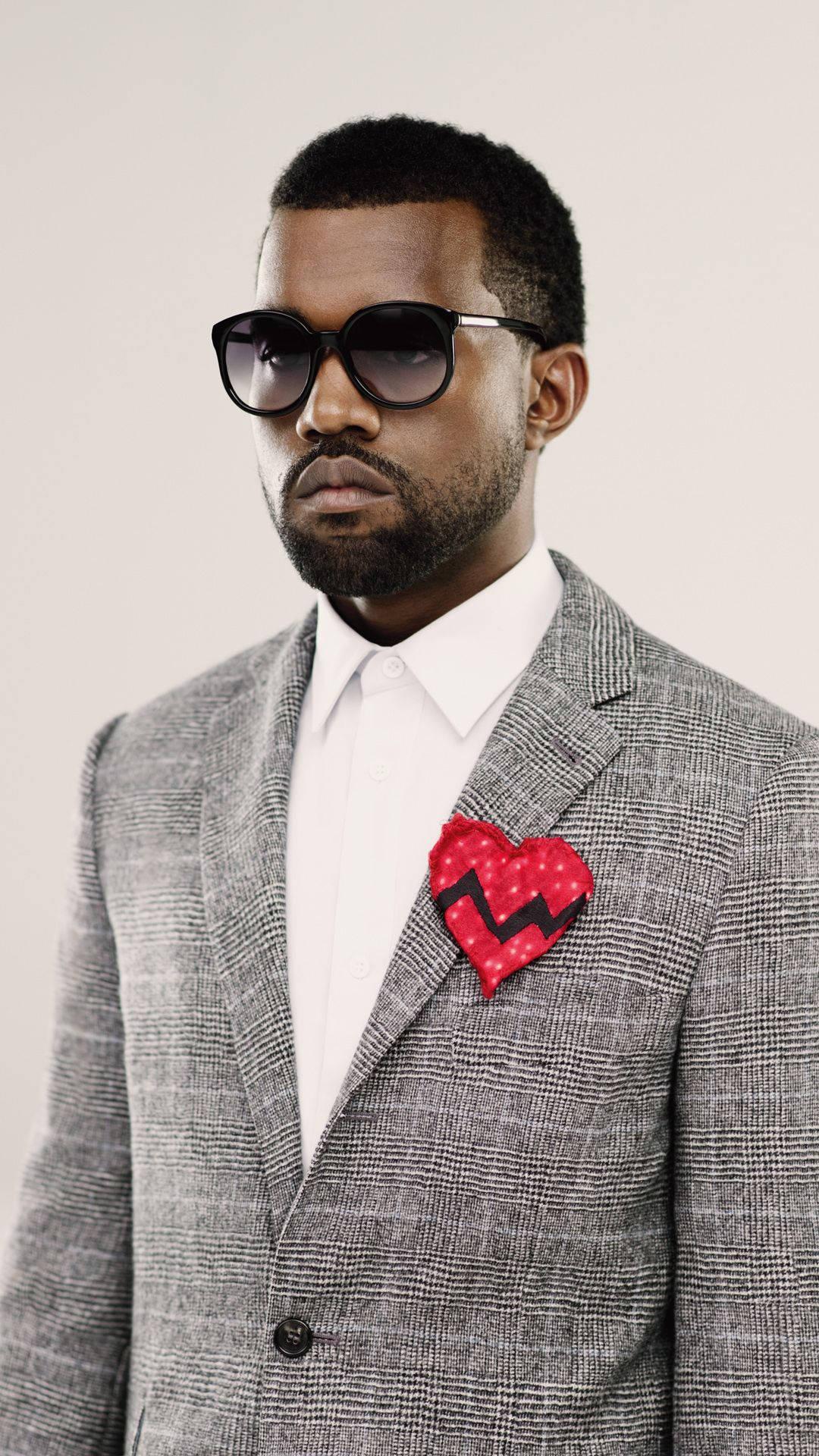Kanye West 桌布 壁紙 — 一位時尚男士穿著灰色格紋西裝和太陽眼鏡，在純色背景前佩戴紅色心形口袋巾。