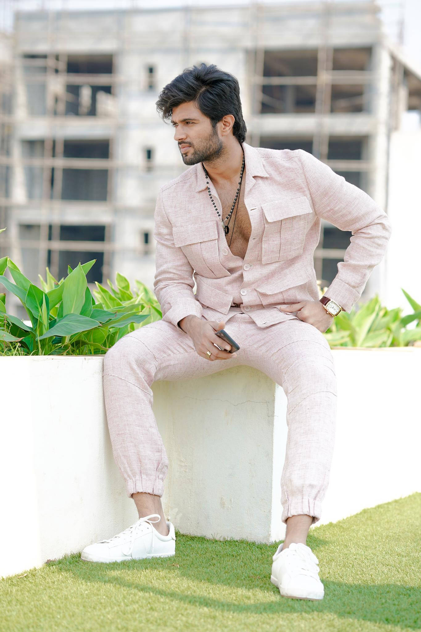vijay devarakonda 4K 桌布 壁紙 — 一位時尚男士穿著粉紅色亞麻西裝，隨意地坐在白色窗台上，身後有綠色植物和建築工地。