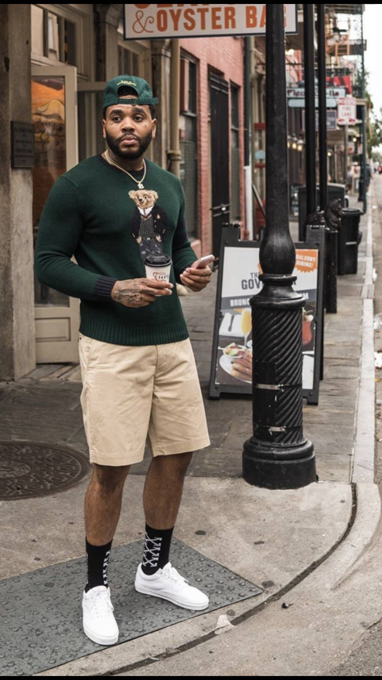 Kevin Gates 桌布 壁紙 — 一位時尚男士穿著綠色毛衣和米色短褲，站在城市人行道上，拿著咖啡和手機。