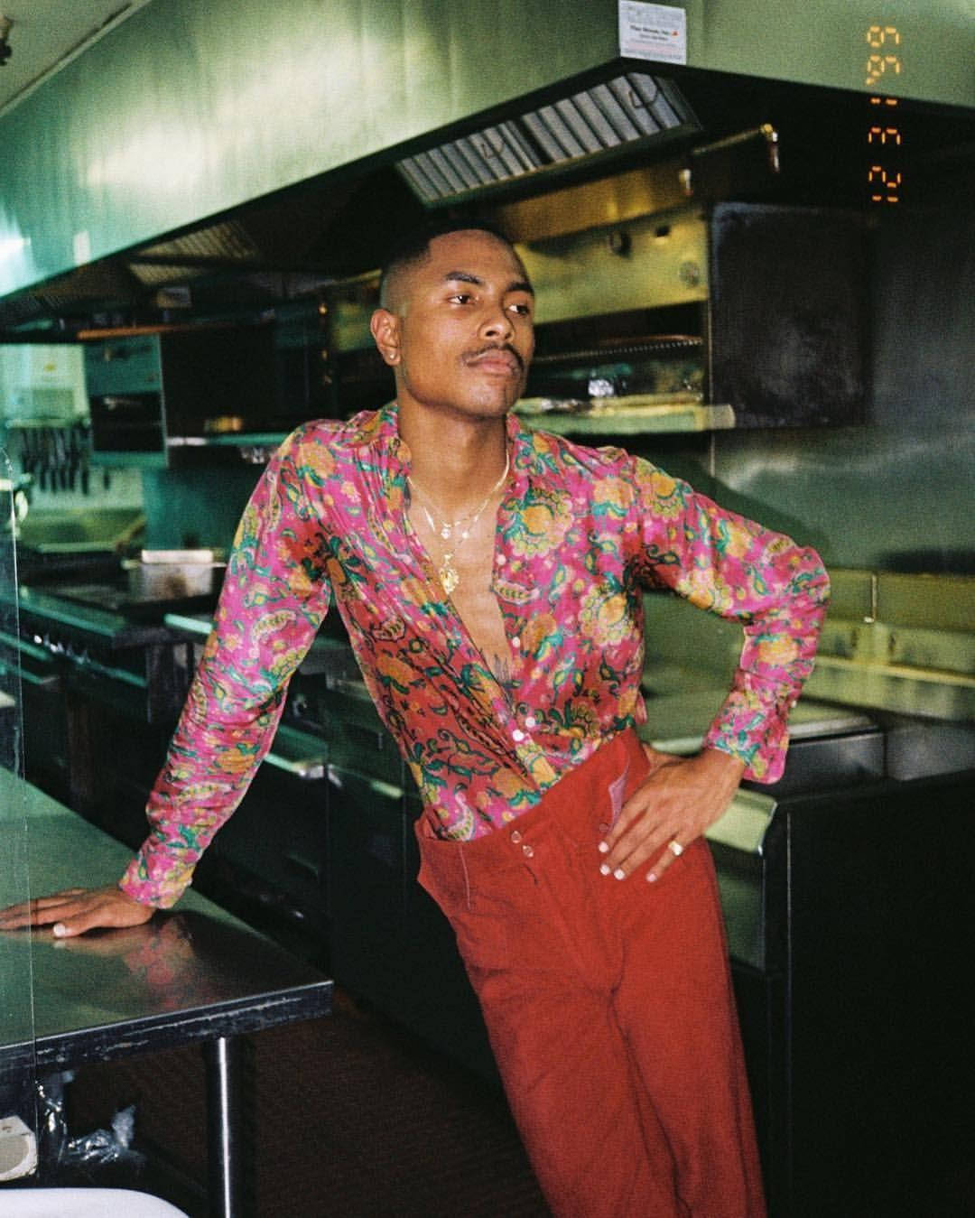 Steve Lacy 桌布 壁紙 — 一位時尚男士穿著鮮豔花卉襯衫和紅色褲子，自信地靠在復古廚房的鋼製檯面上。
