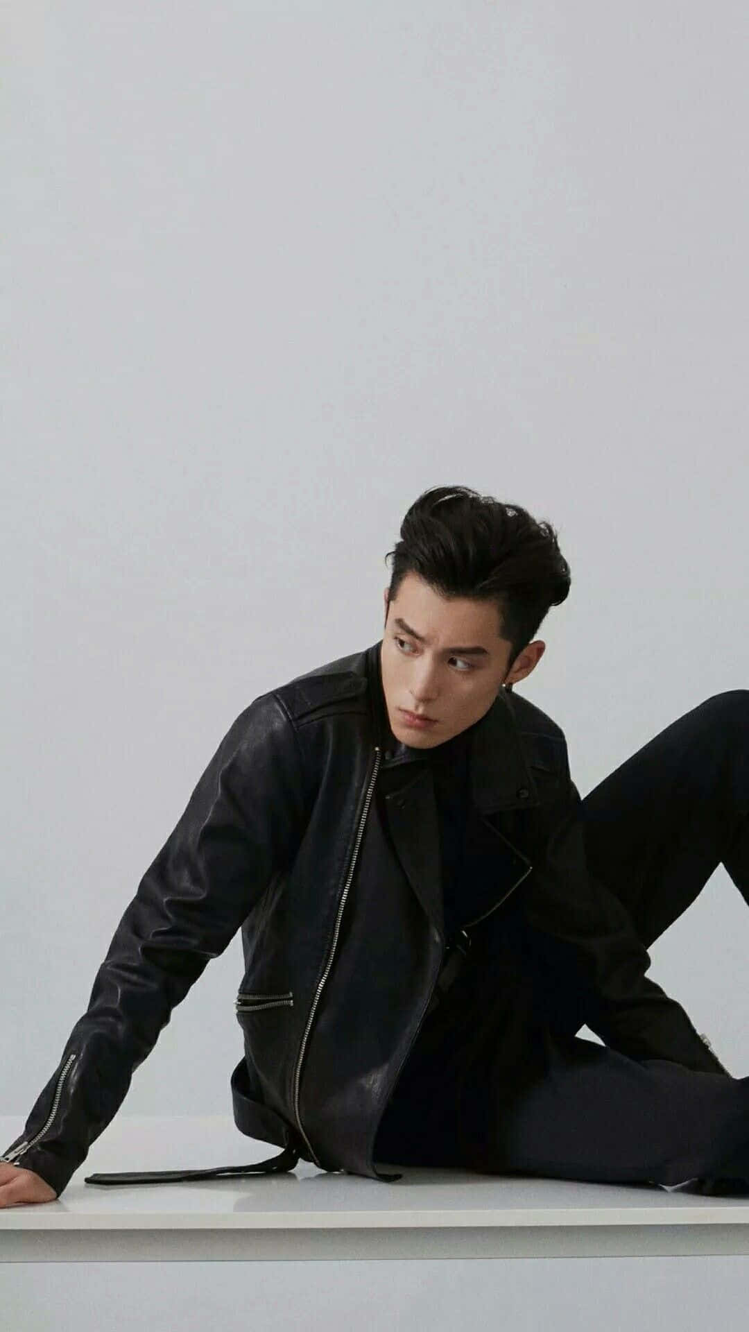 Dylan Wang 桌布 壁紙 — 一位時尚男士穿著黑色皮夾克，自信地坐在白色表面上，背景是純淺灰色。