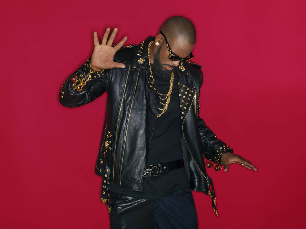 r kelly 桌布 壁紙 — 一位時尚男士穿著黑色皮夾克，飾有金鏈和鉚釘，在鮮紅背景前自信地擺姿勢。