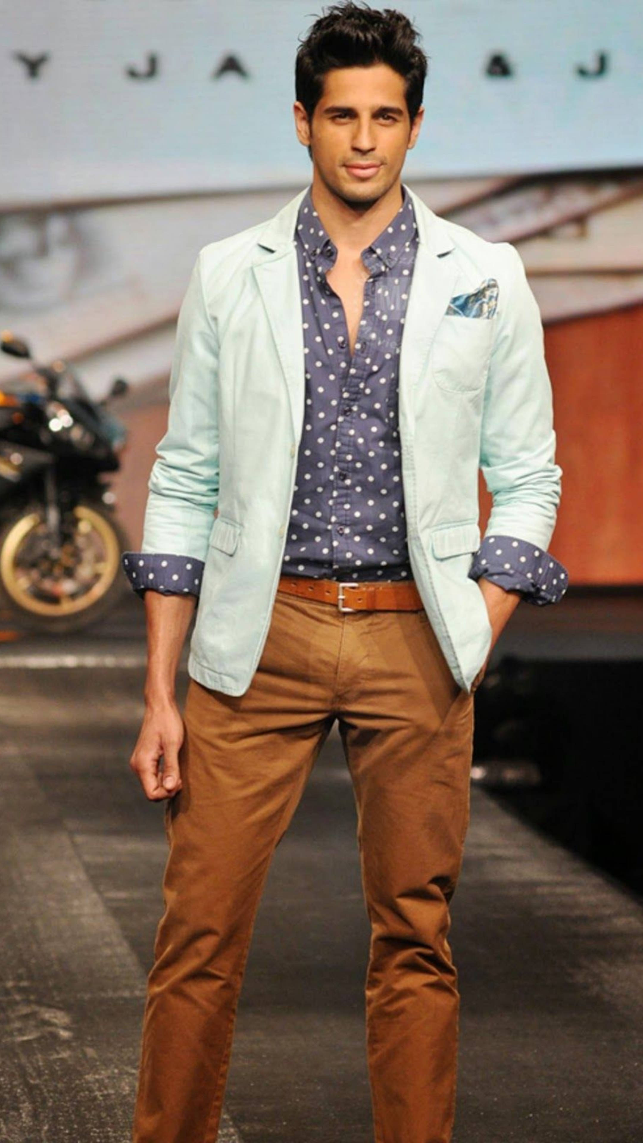 Sidharth Malhotra 桌布 壁紙 — 一位時尚男士自信地走在伸展台上，身穿淺藍色西裝外套，內搭圓點襯衫和棕色長褲。