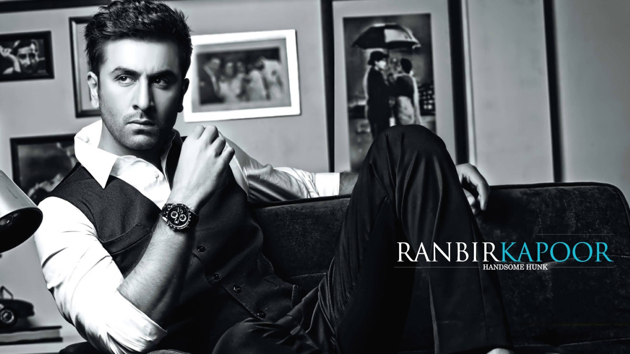 Ranbir Kapoor 桌布 壁紙 — 一位時尚男士自信地躺臥在沙發上，身處一個以裱框照片和復古裝飾點綴的單色房間。