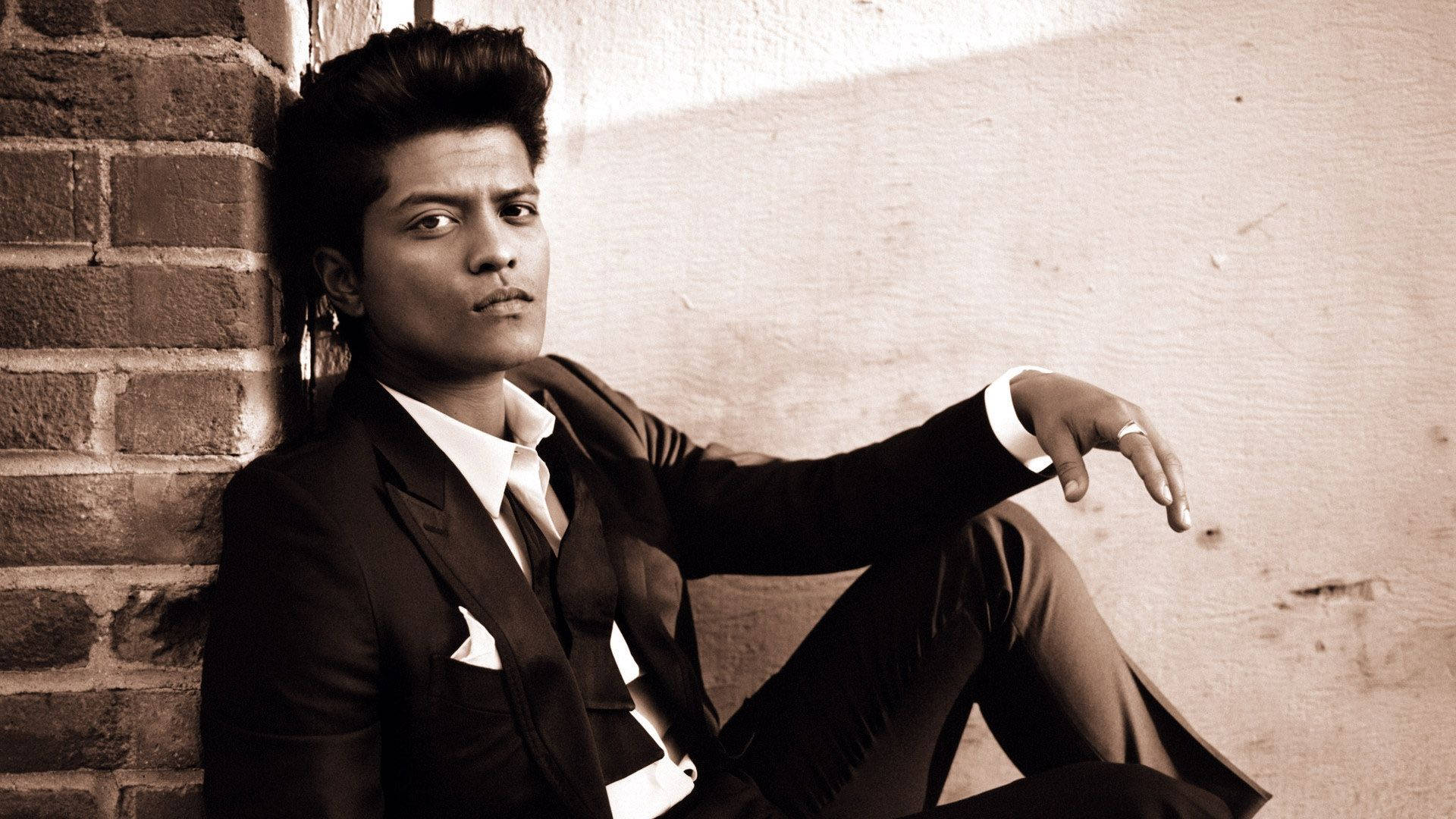 Bruno Mars 桌布 壁紙 — 一位時尚男士身穿俐落西裝，在溫暖的棕褐色燈光下，以自信的目光靠在磚牆上。