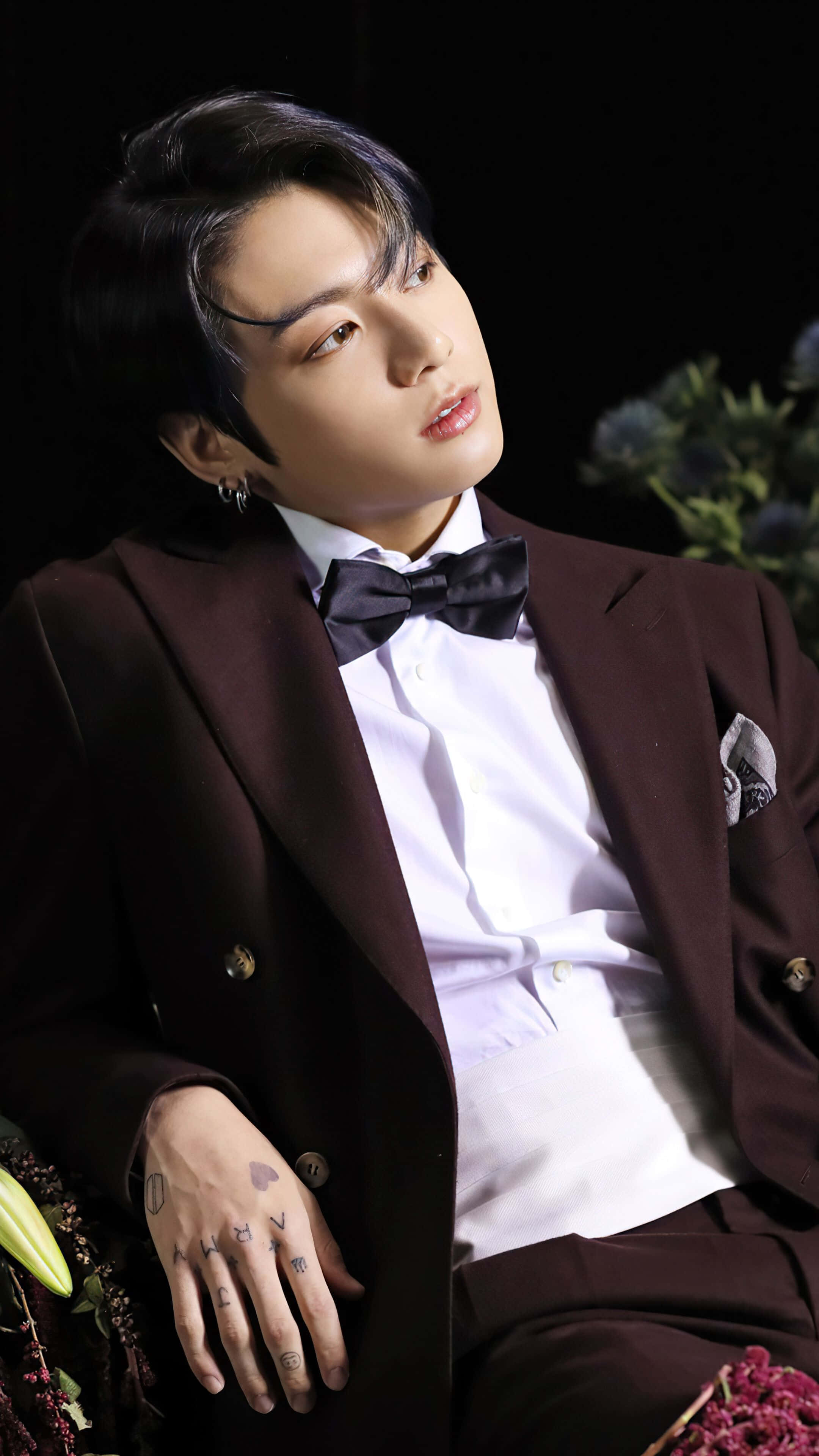 Jungkook 桌布 壁紙 — 一位時尚男士身穿栗色西裝和黑色領結，在帶有花卉裝飾的深色背景前擺姿勢。