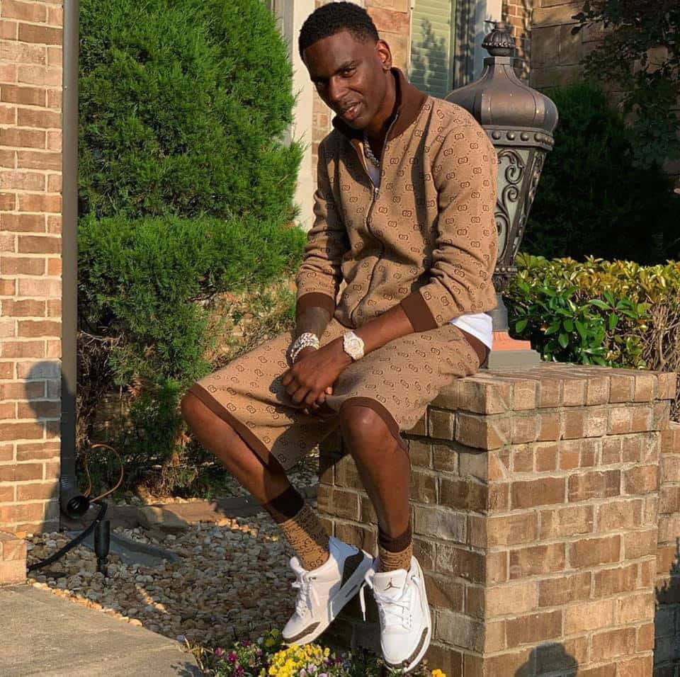 Young Dolph 桌布 壁紙 — 一位時尚男士身穿棕色圖案運動服，自信地坐在戶外的磚柱上，背景有綠色灌木和房屋。