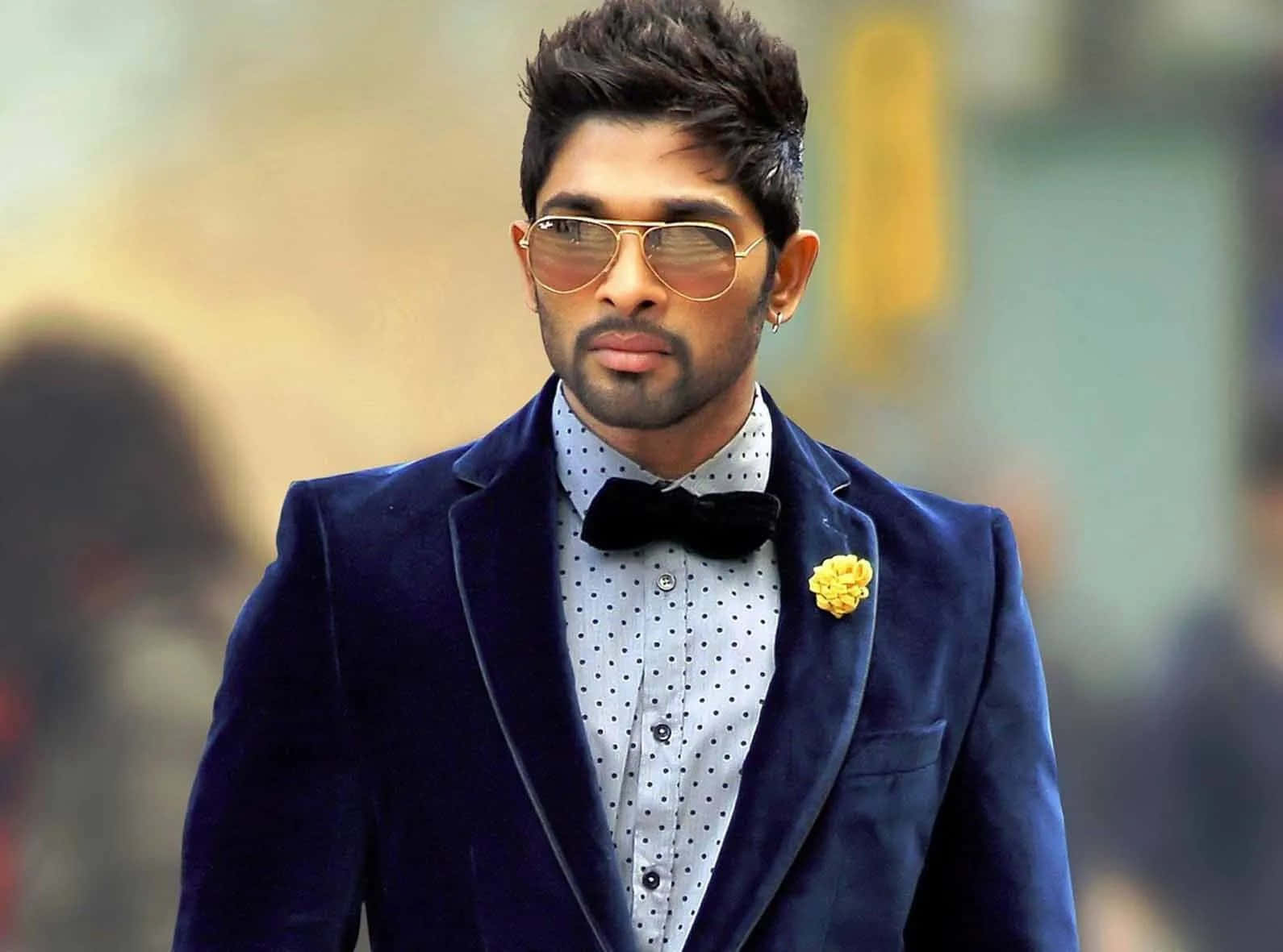 Allu Arjun 桌布 壁紙 — 一位時尚男士身穿海軍藍天鵝絨西裝，佩戴黃色花朵胸花和飛行員太陽眼鏡，自信地行走。