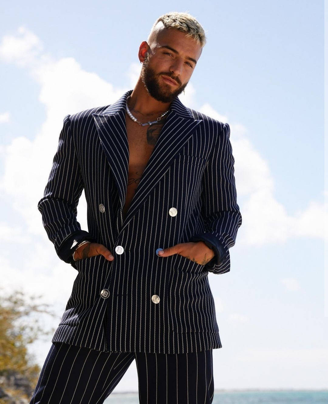Maluma 桌布 壁紙 — 一位時尚男士身穿海軍藍細條紋西裝，雙手插袋，自信地站在戶外，背景是明亮的藍天。