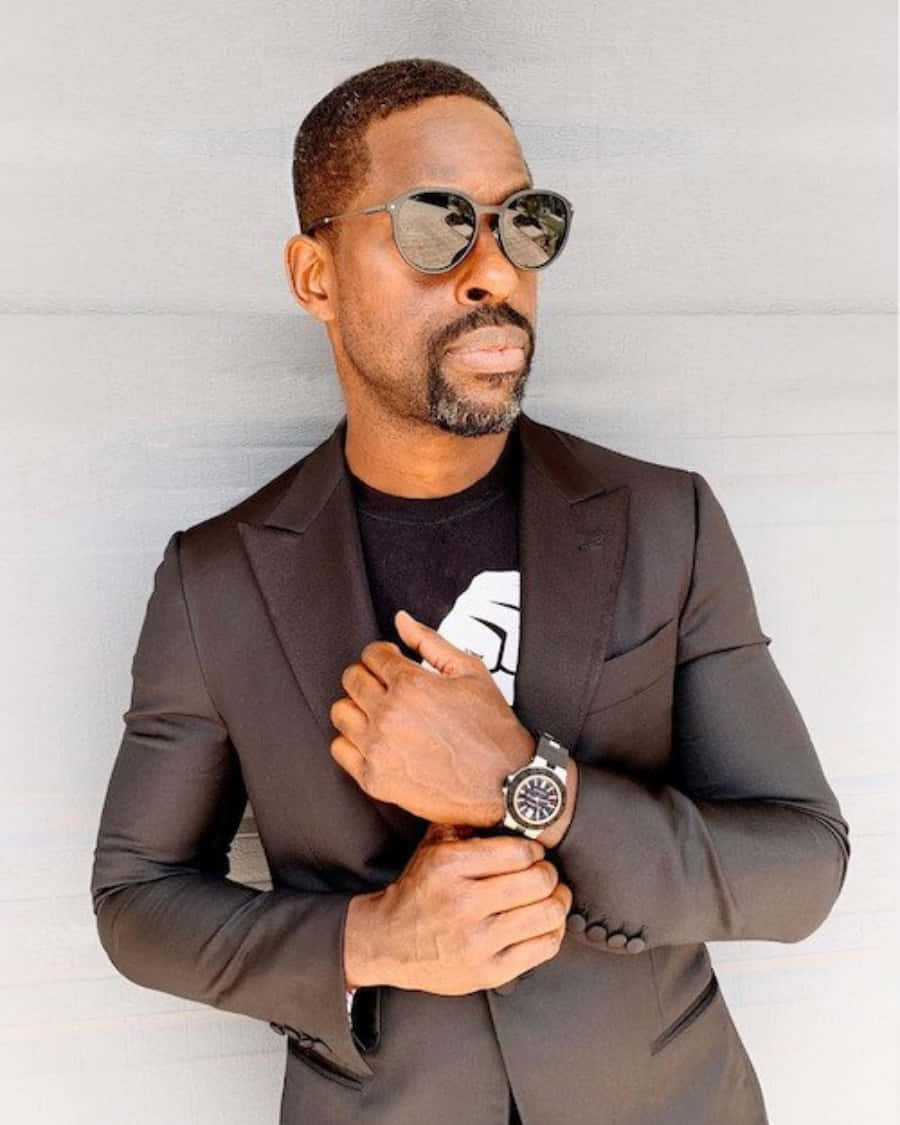 Sterling K Brown 桌布 壁紙 — 一位時尚男士身穿深色西裝和太陽眼鏡，自信地站在純白牆壁前，手腕上戴著手錶。