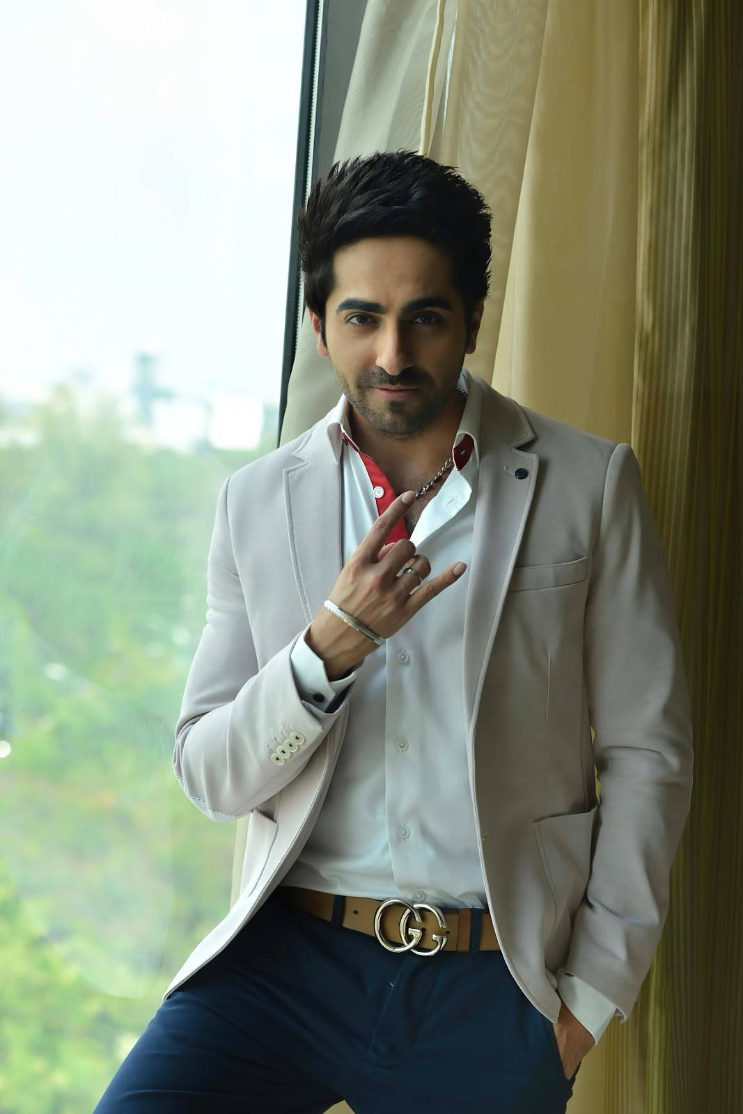 ayushmann khurrana 桌布 壁紙 — 一位時尚男士身穿淺色西裝外套和紅色襯衫，自信地在窗邊擺姿勢，窗外有綠植。
