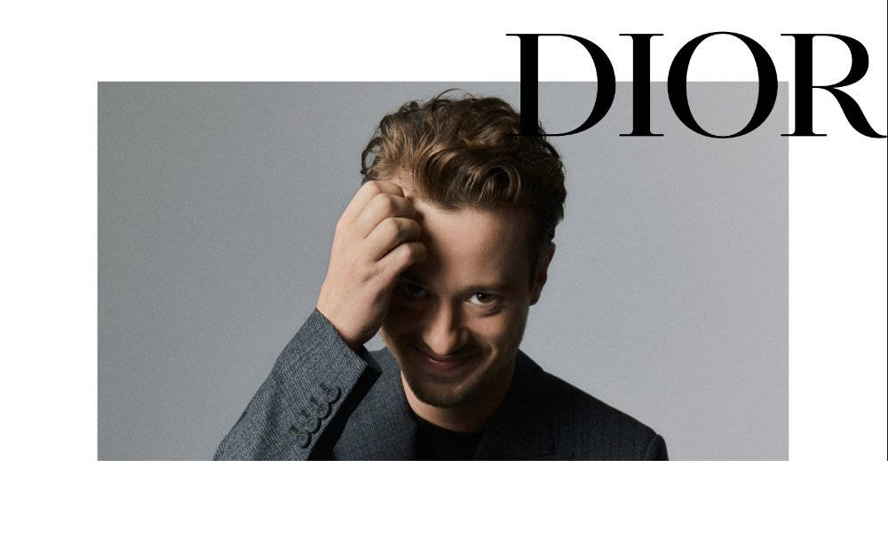 身穿灰色西裝的男士在灰色背景上微笑，手放在頭上，帶有DIOR文字。 — Dior 動態 壁紙