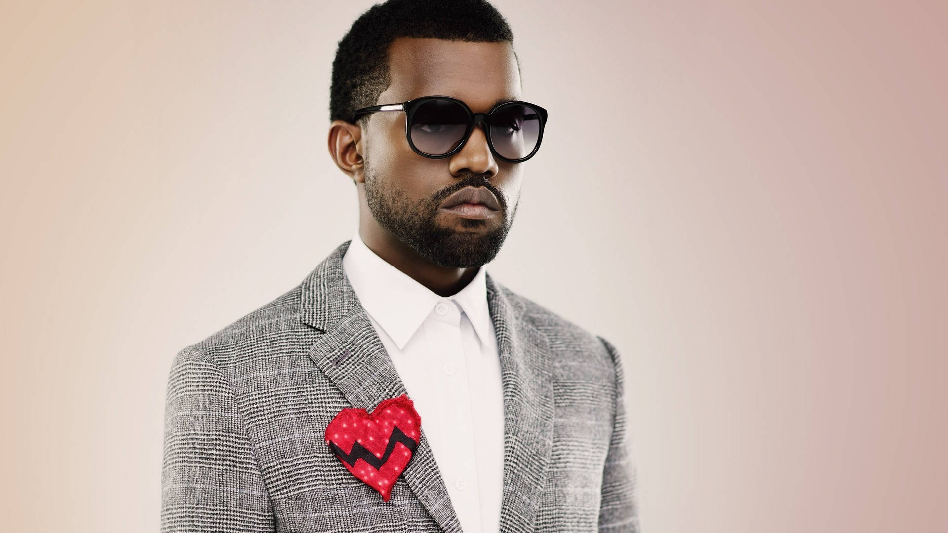 Kanye 桌布 壁紙 — 一位時尚男士身穿灰色西裝和太陽眼鏡，在柔和的粉紅色背景上佩戴紅色破碎心形別針。