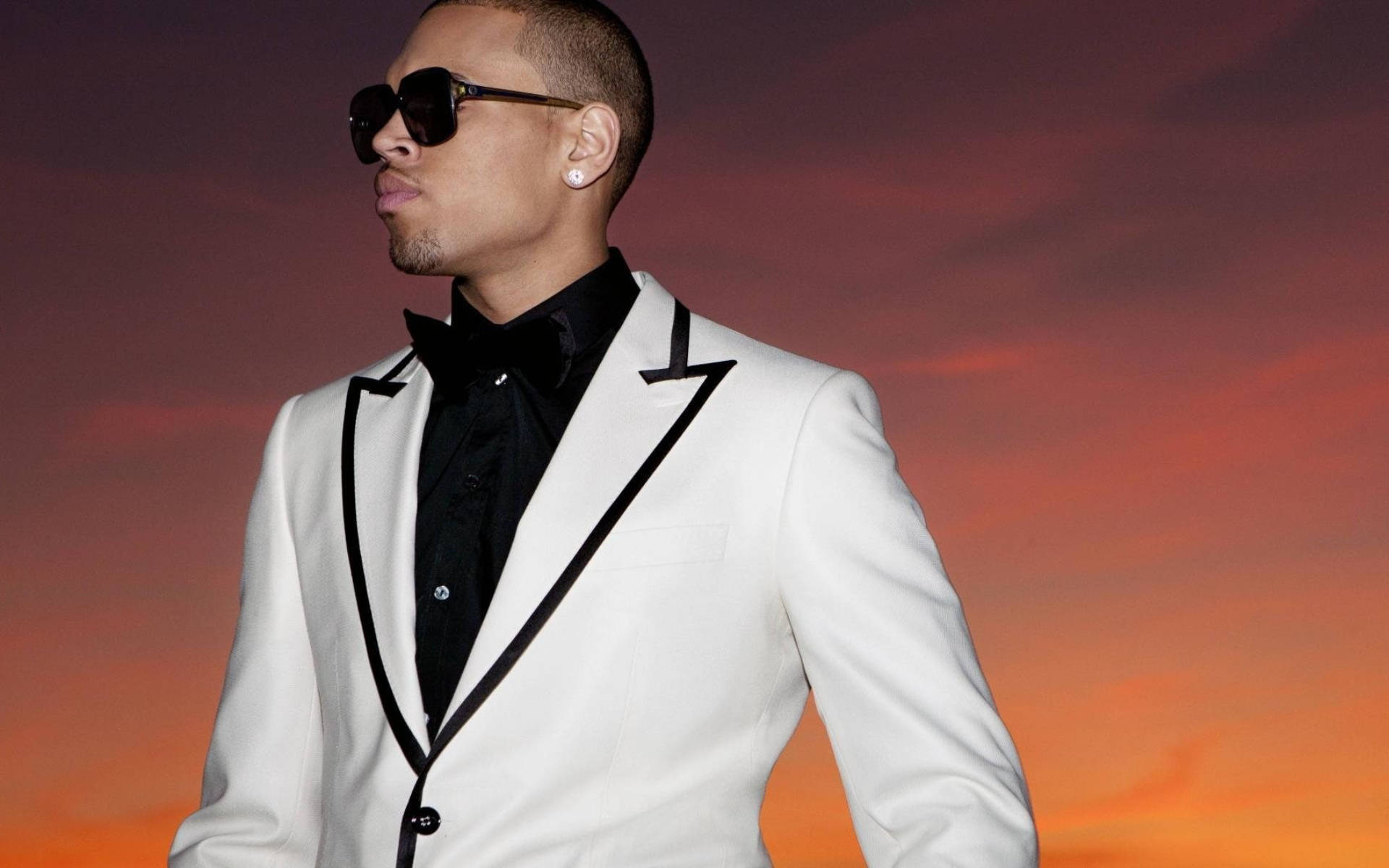 Chris Brown 桌布 壁紙 — 一位時尚男士身穿白色燕尾服配黑色領，自信地站在充滿活力的日落天空前。