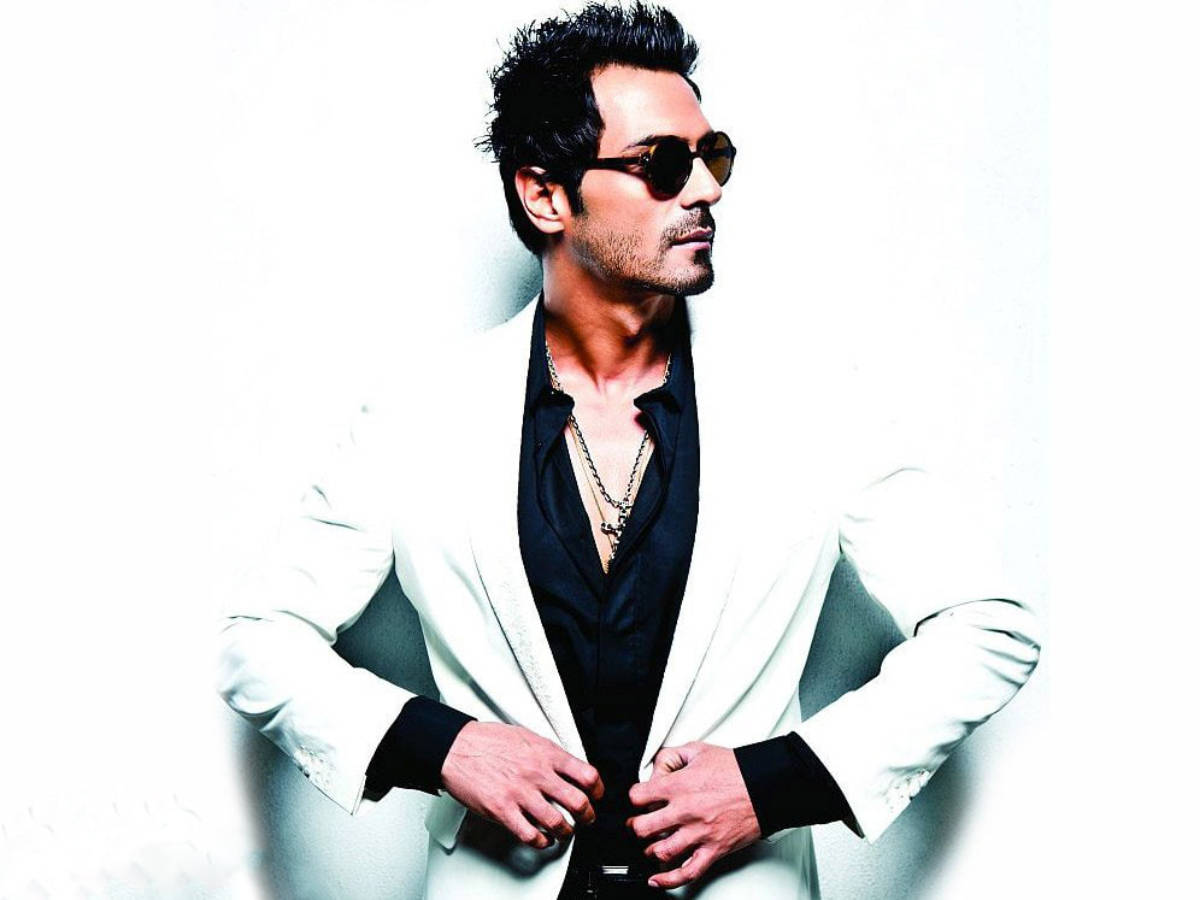 Arjun Rampal 桌布 壁紙 — 一位時尚男士身穿白色西裝外套和黑色襯衫，戴著太陽眼鏡，在純白背景前自信地擺姿勢。