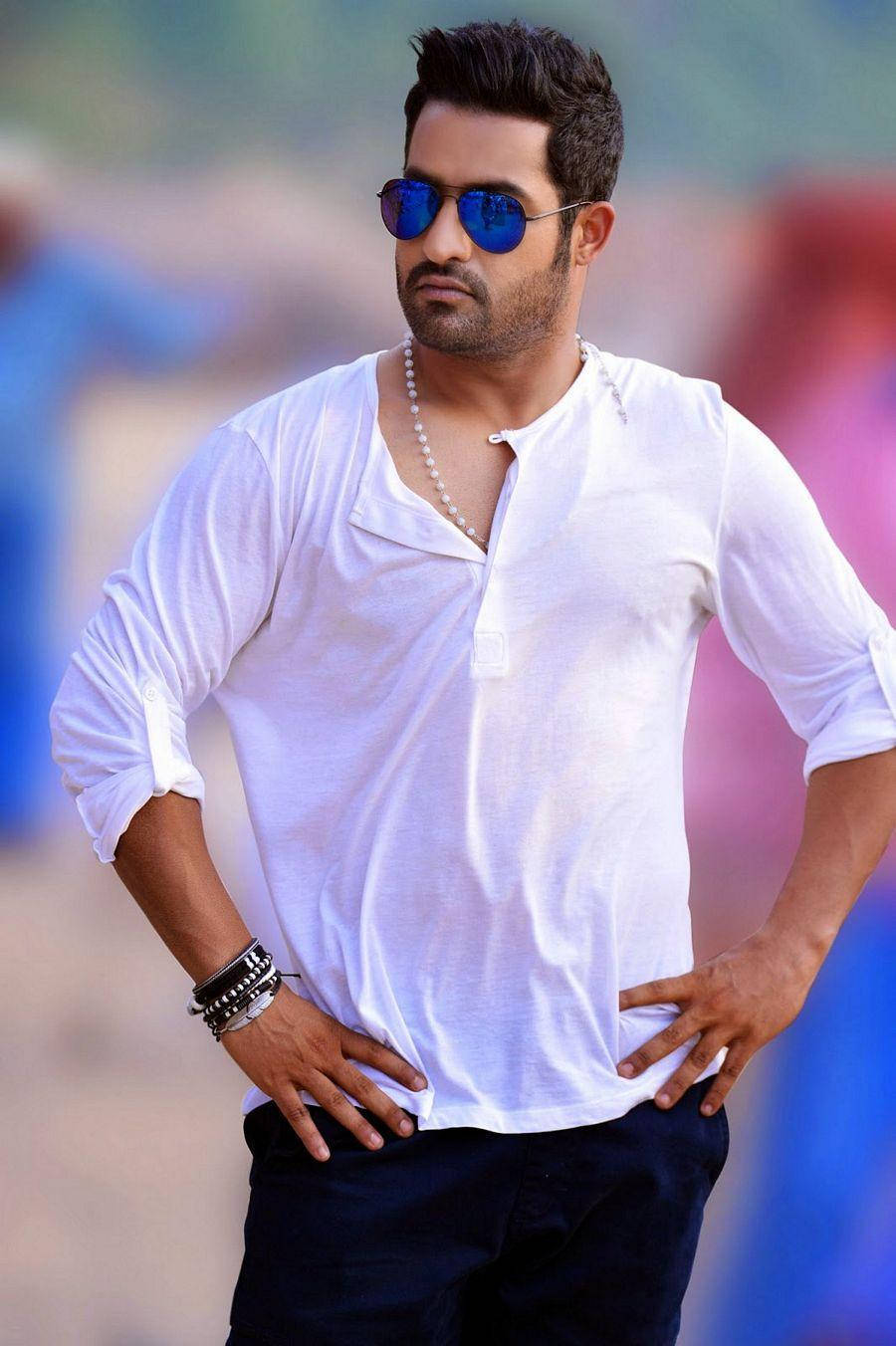 jr ntr 桌布 壁紙 — 一位時尚男士身穿白襯衫和藍色太陽眼鏡，雙手叉腰，自信地站在色彩繽紛的模糊背景前。