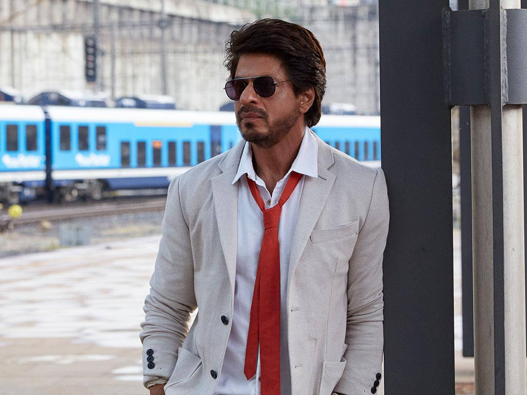 Shahrukh 桌布 壁紙 — 一位時尚男士身穿米色西裝和紅色領帶，在火車站靠著柱子，一列藍色火車駛過。