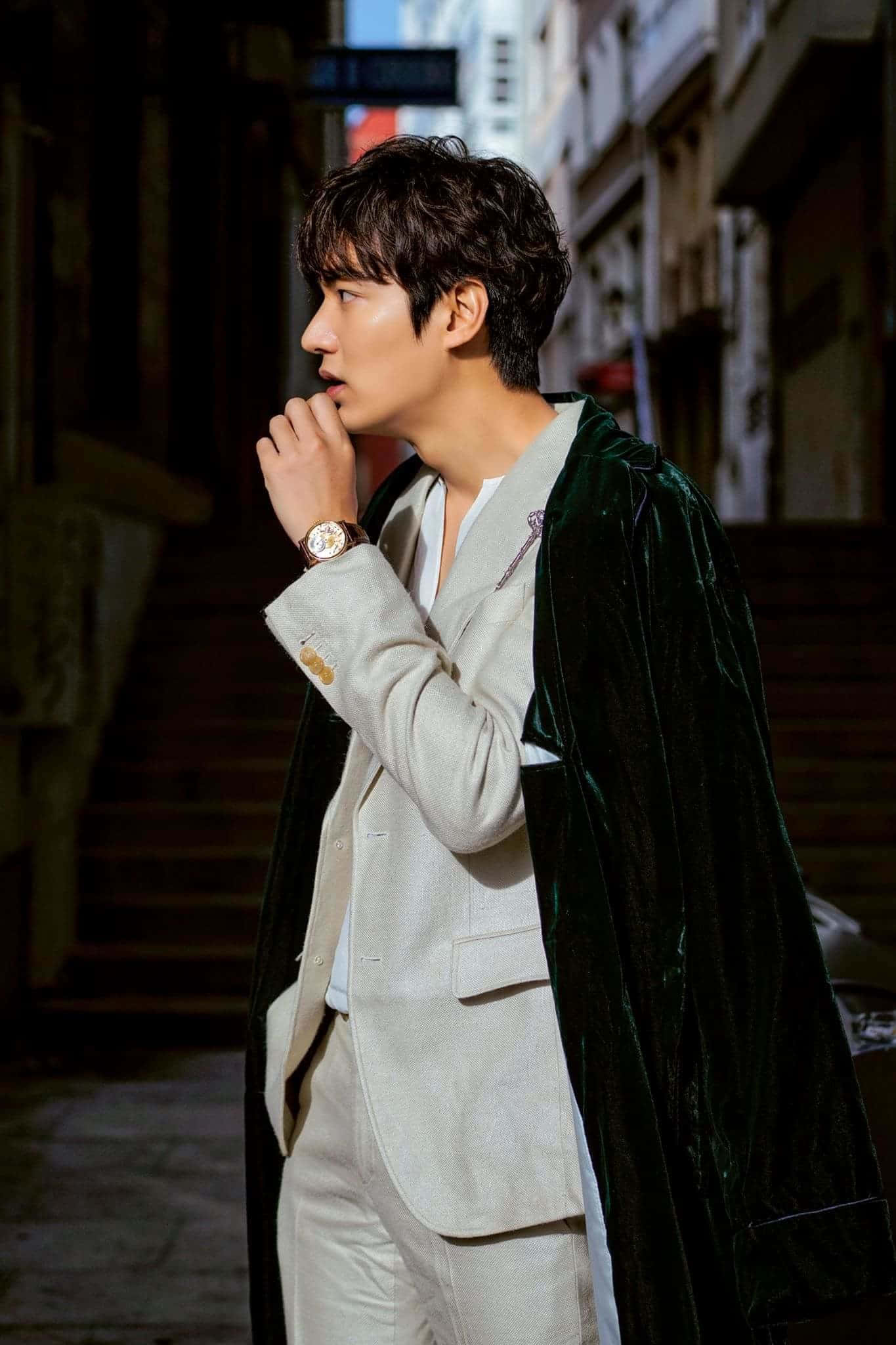 Lee Min Ho 桌布 壁紙 — 一位時尚男士身穿米色西裝和綠色天鵝絨斗篷，站在陽光普照的城市小巷中，手靠近嘴巴，作沉思狀。