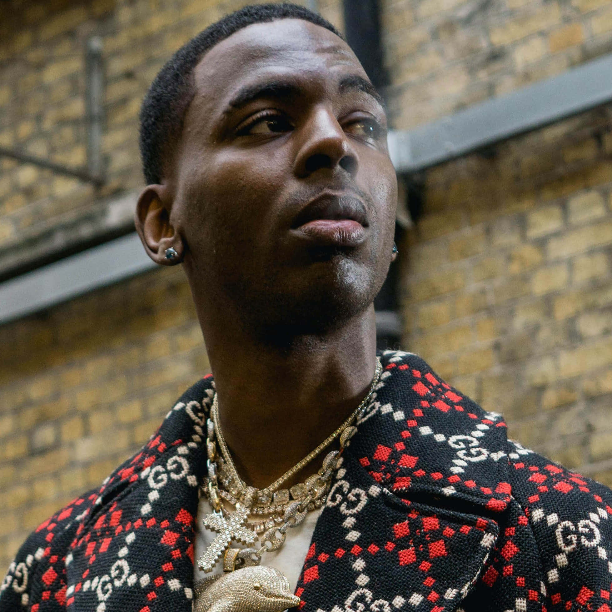 Young Dolph 桌布 壁紙 — 一位時尚男士身穿紅黑圖案Gucci外套和金色鏈條，在磚牆背景下向上凝視。