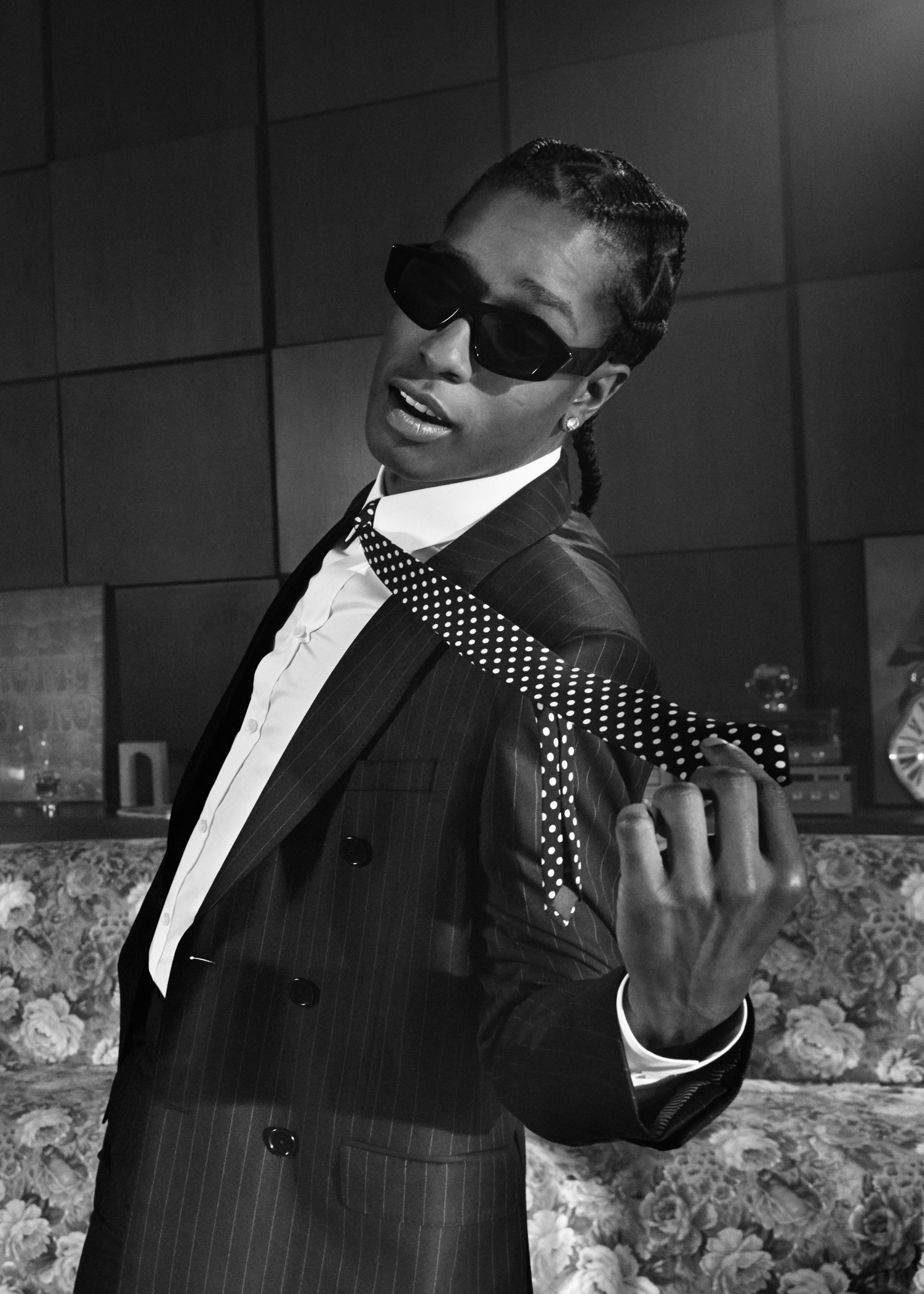 rocky 桌布 壁紙 — 一位時尚男士身穿細條紋西裝和波點領帶，在單色房間內搭配花卉軟墊，自信地戴著太陽眼鏡擺姿勢。