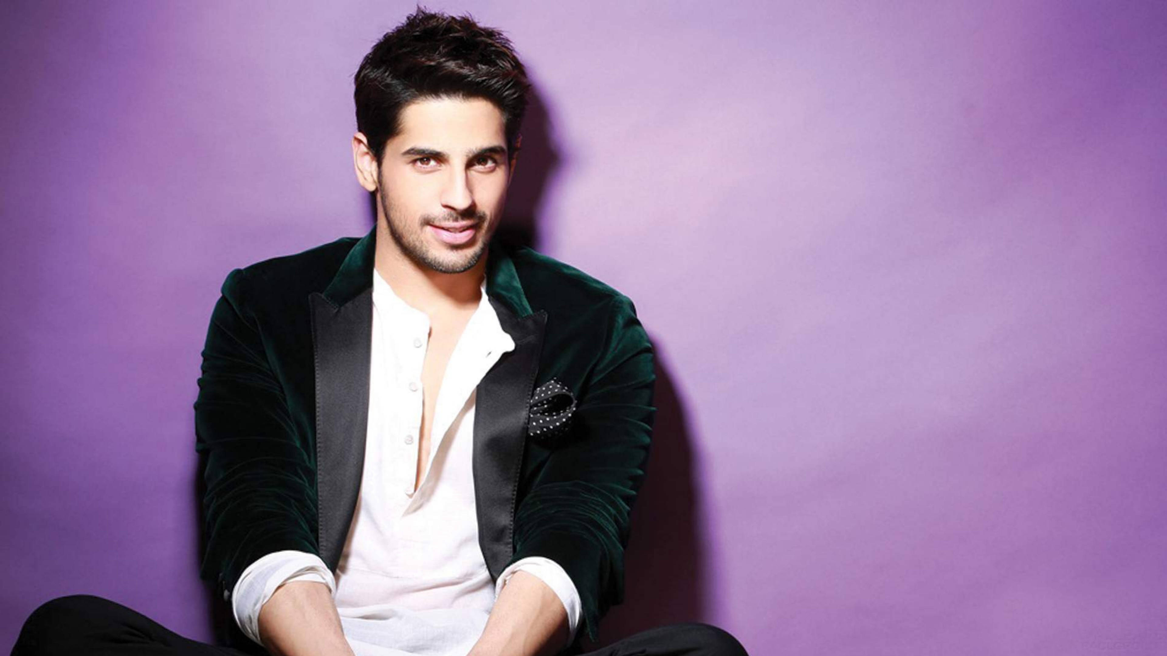 Sidharth Malhotra 桌布 壁紙 — 一位時尚男士身穿綠色天鵝絨西裝外套和白色襯衫，在純紫色攝影棚背景前自信地擺姿勢。