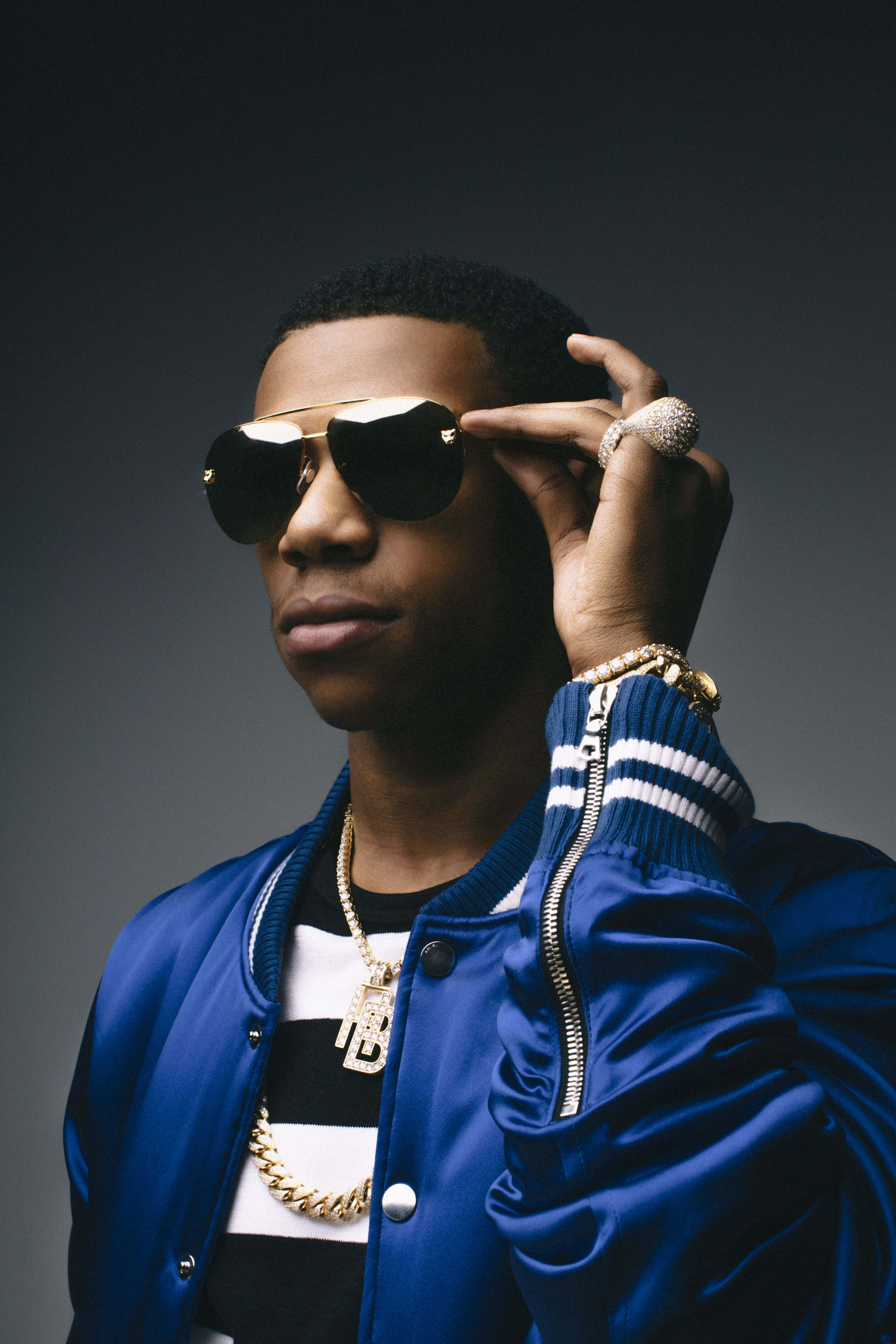 Rappers 桌布 壁紙 — 一位時尚男士身穿藍色轟炸機夾克，佩戴金鍊和鑽石戒指，調整他的飛行員太陽眼鏡。