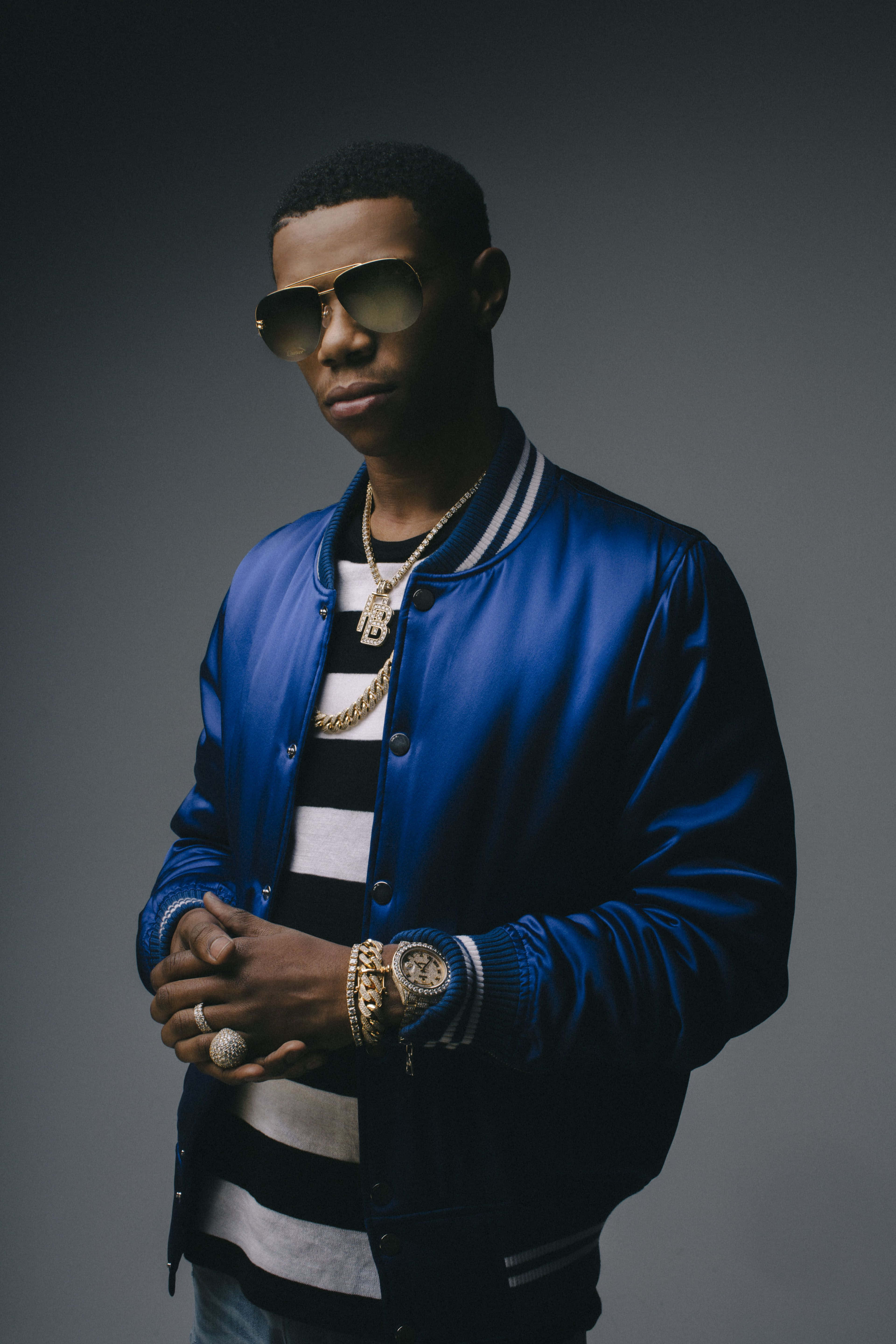 Rappers 桌布 壁紙 — 一位時尚男士身穿藍色飛行員夾克和金色鏈條，在深灰色工作室背景前自信地擺姿勢。