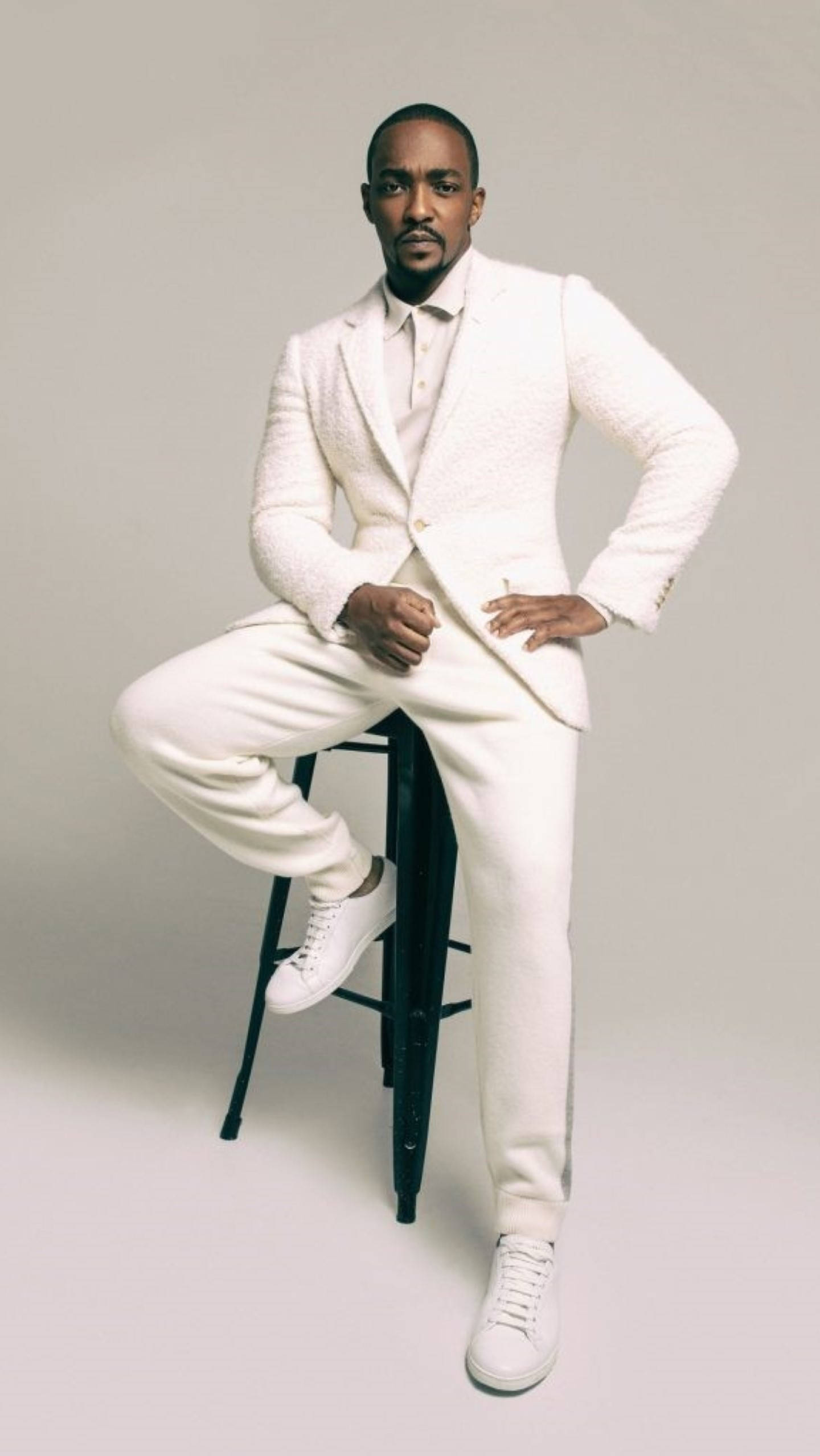 Anthony Mackie 桌布 壁紙 — 一位時尚男士身穿質感白色西裝和運動鞋，自信地坐在黑色凳子上，背景是純灰色攝影棚。
