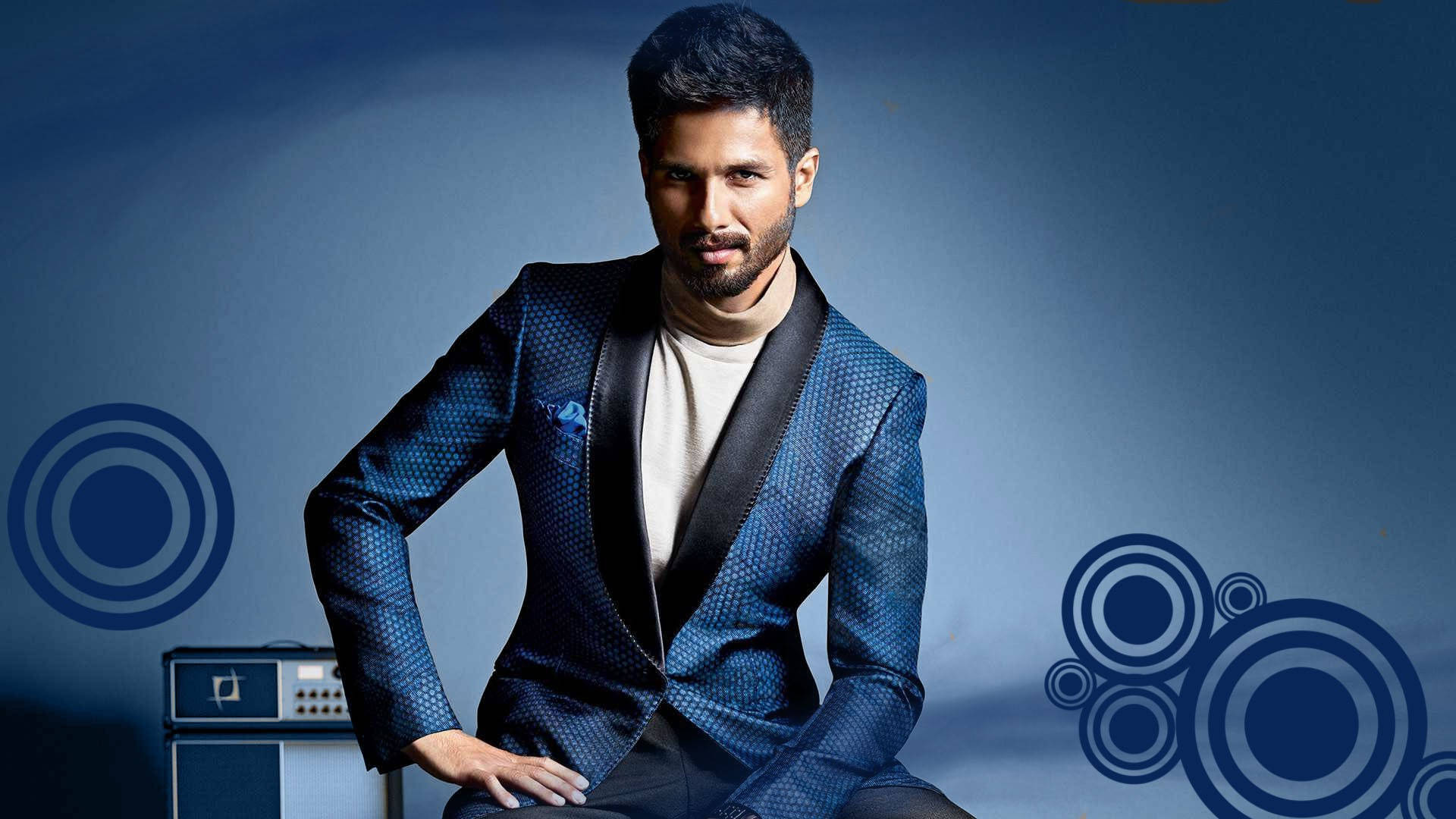 Shahid Kapoor 桌布 壁紙 — 一位時尚男士身穿質感藍色西裝，在帶有圓形圖形元素的漸變藍色背景前自信地擺姿勢。