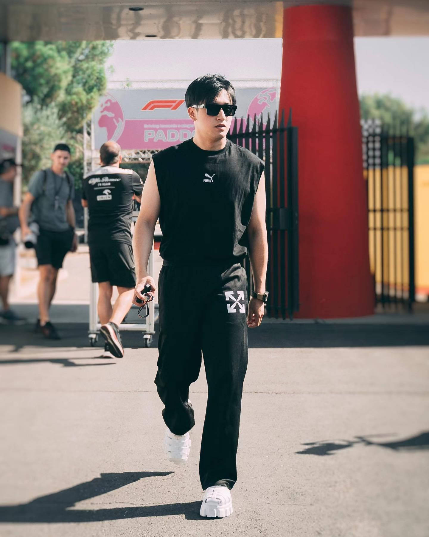 Sunglasses 桌布 壁紙 — 一位時尚男士身穿黑色無袖上衣和長褲，搭配白色運動鞋，自信地走過帶有F1品牌標誌的陽光戶外場地。