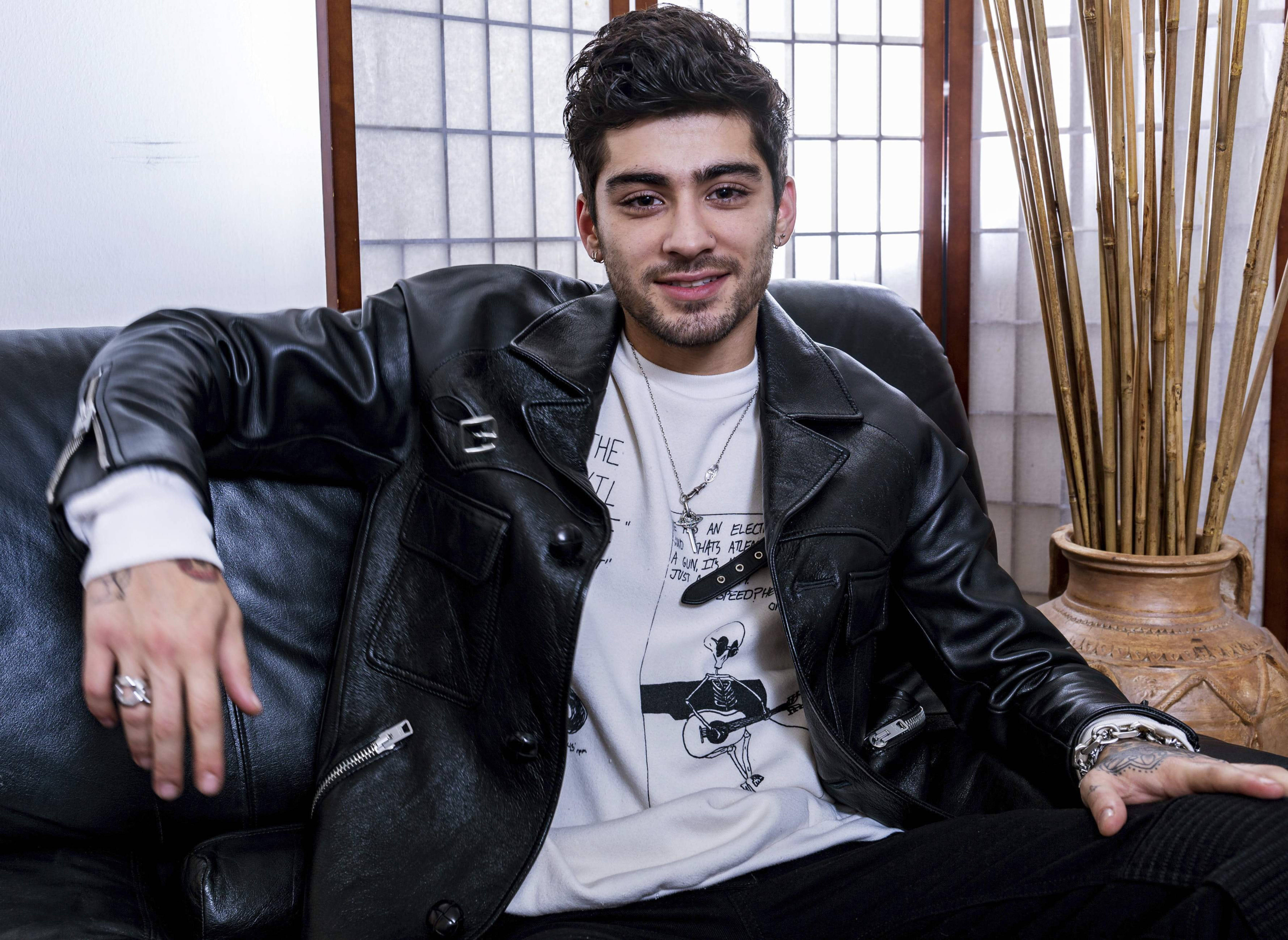 zayn 桌布 壁紙 — 一位時尚男士身穿黑色皮夾克，在深色沙發上放鬆，旁邊放著竹花瓶，面露微笑。