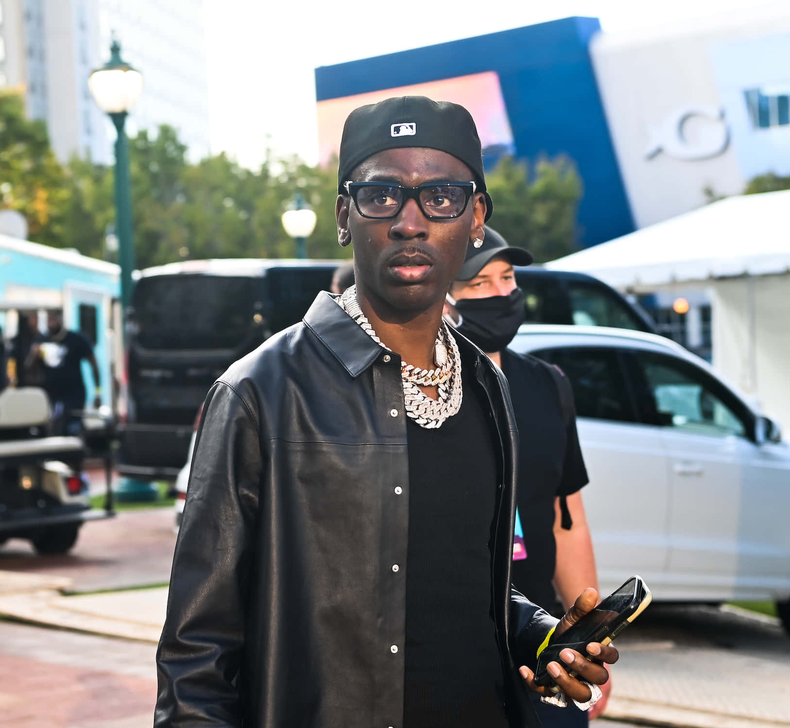 Young Dolph 桌布 壁紙 — 一位時尚男士身穿黑色皮夾克和帽子，自信地站在戶外，手持電話，背景是城市車輛和建築物。