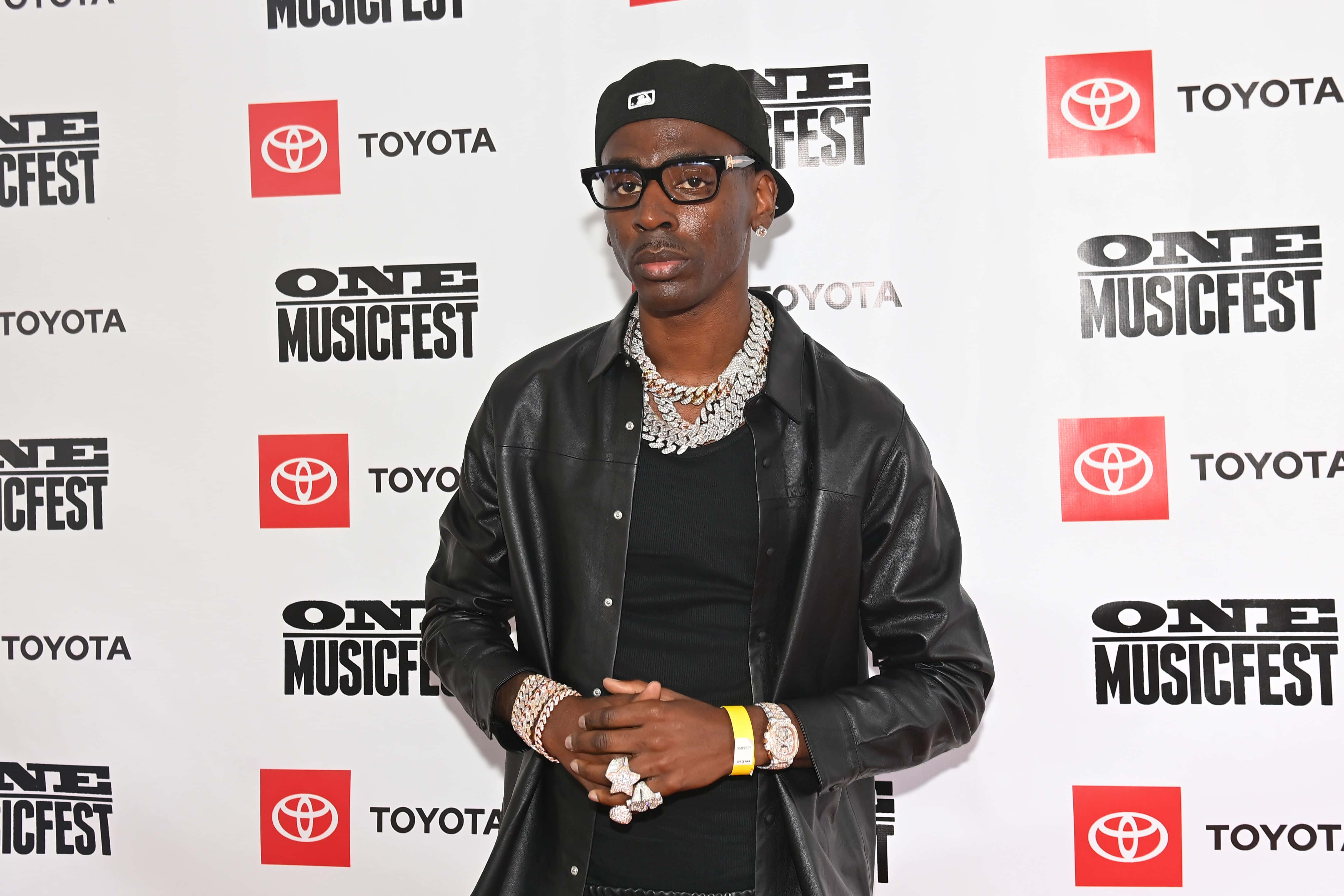 Young Dolph 桌布 壁紙 — 一位時尚男士身穿黑色皮夾克和帽子，在背後有豐田品牌的One Music Fest活動中自信地擺姿勢。