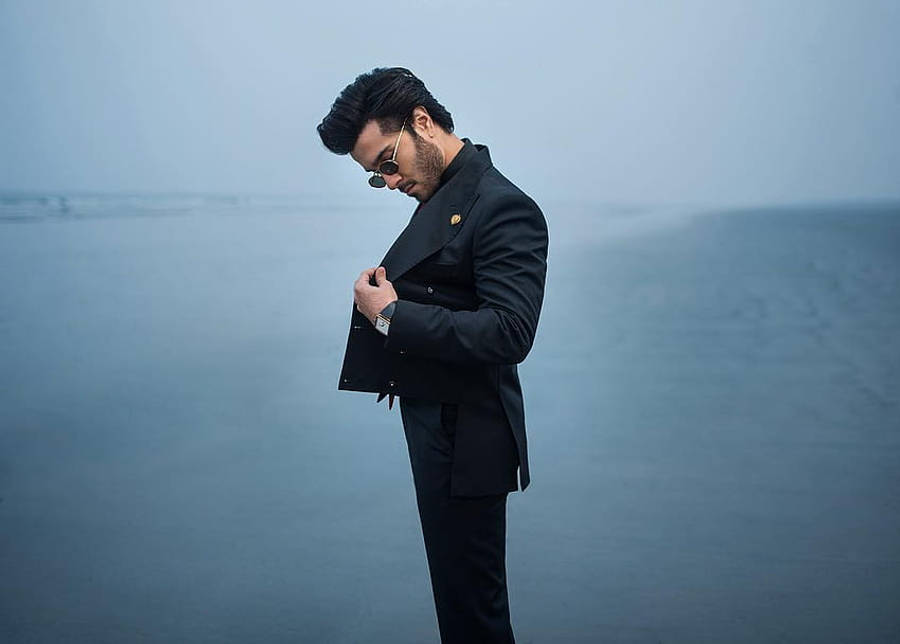 Feroz Khan 桌布 壁紙 — 一位時尚男士身穿黑色西裝和太陽眼鏡，在霧氣瀰漫的藍色海洋背景下扣上外套鈕扣。