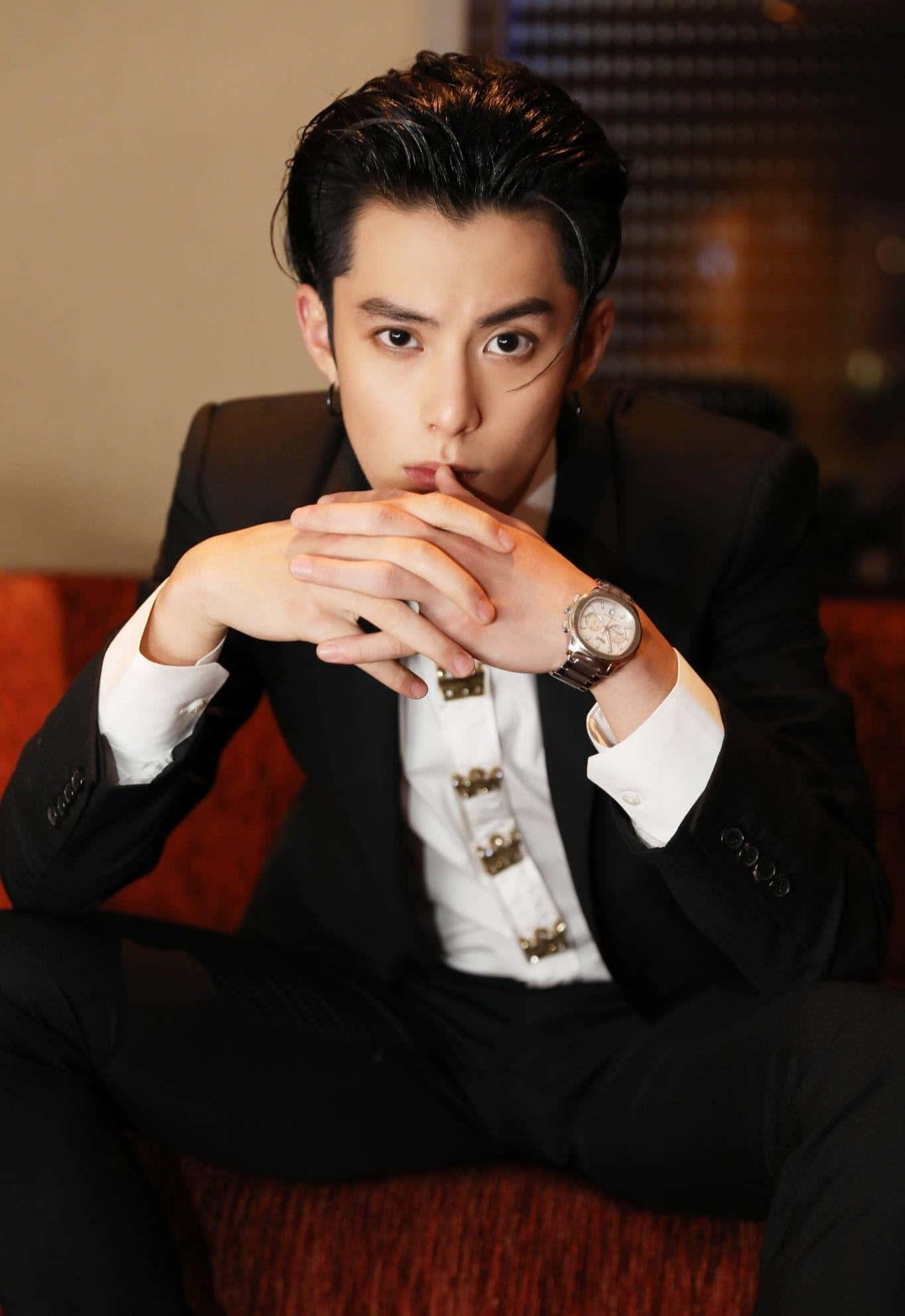 Dylan Wang 桌布 壁紙 — 一位時尚男士身穿黑色西裝和白色襯衫，坐在紅色沙發上，雙手交握，佩戴銀色手錶和耳環。