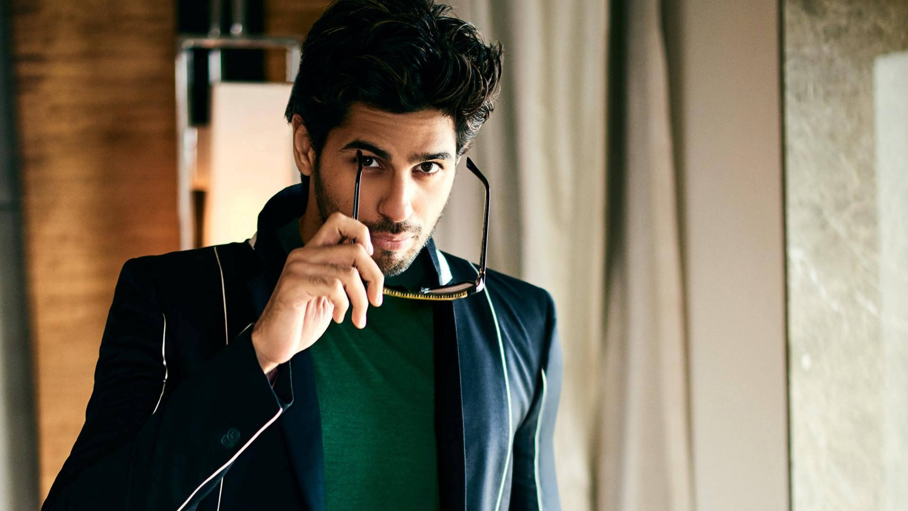 Sidharth Malhotra 桌布 壁紙 — 一位時尚男士身穿黑色西裝外套和綠色襯衫，手持眼鏡框，在溫暖的室內燈光下直視鏡頭。