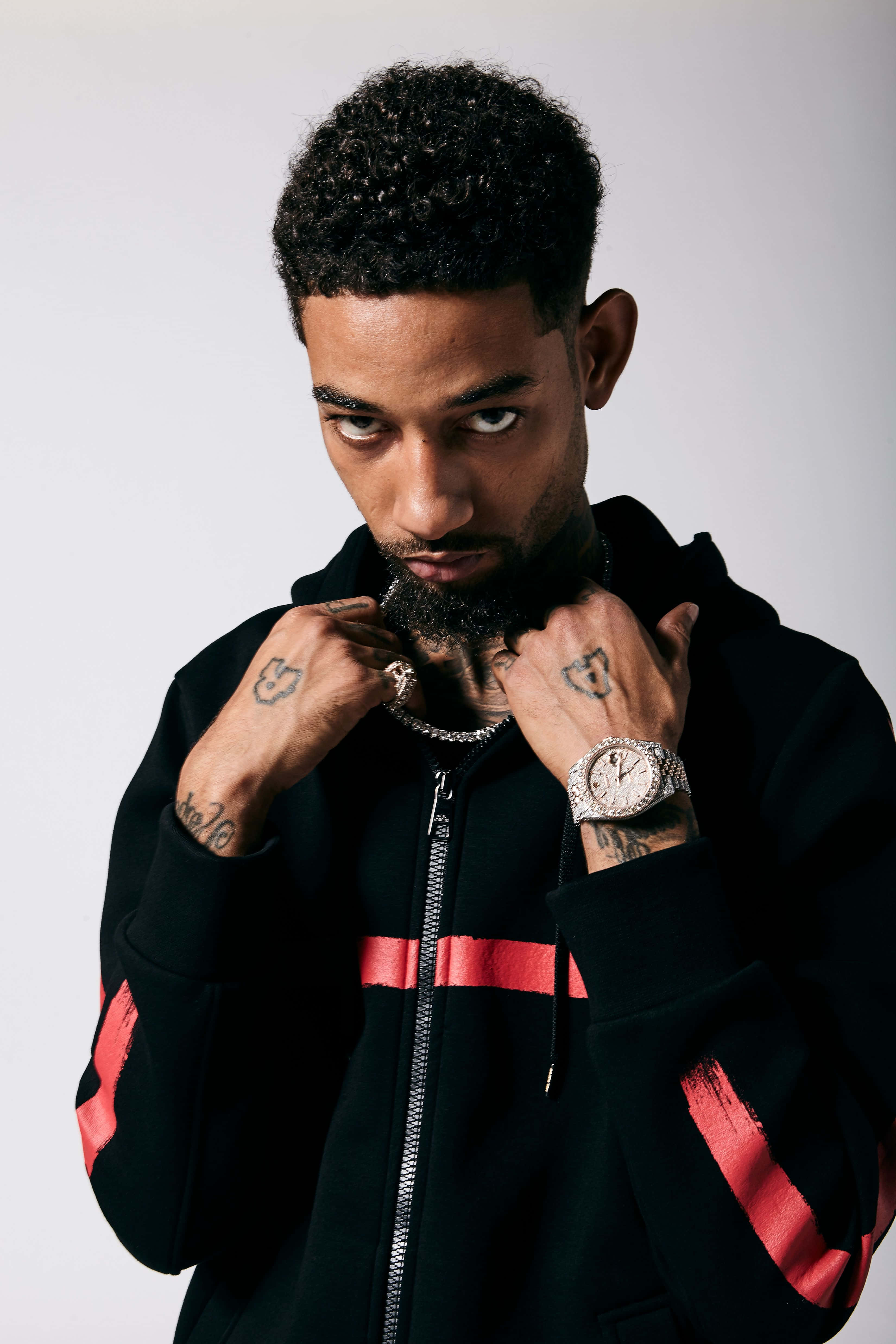 Rappers 桌布 壁紙 — 一位時尚男子，身上有刺青並佩戴鑽石手錶，在純白背景前調整他的黑色紅條連帽衫。