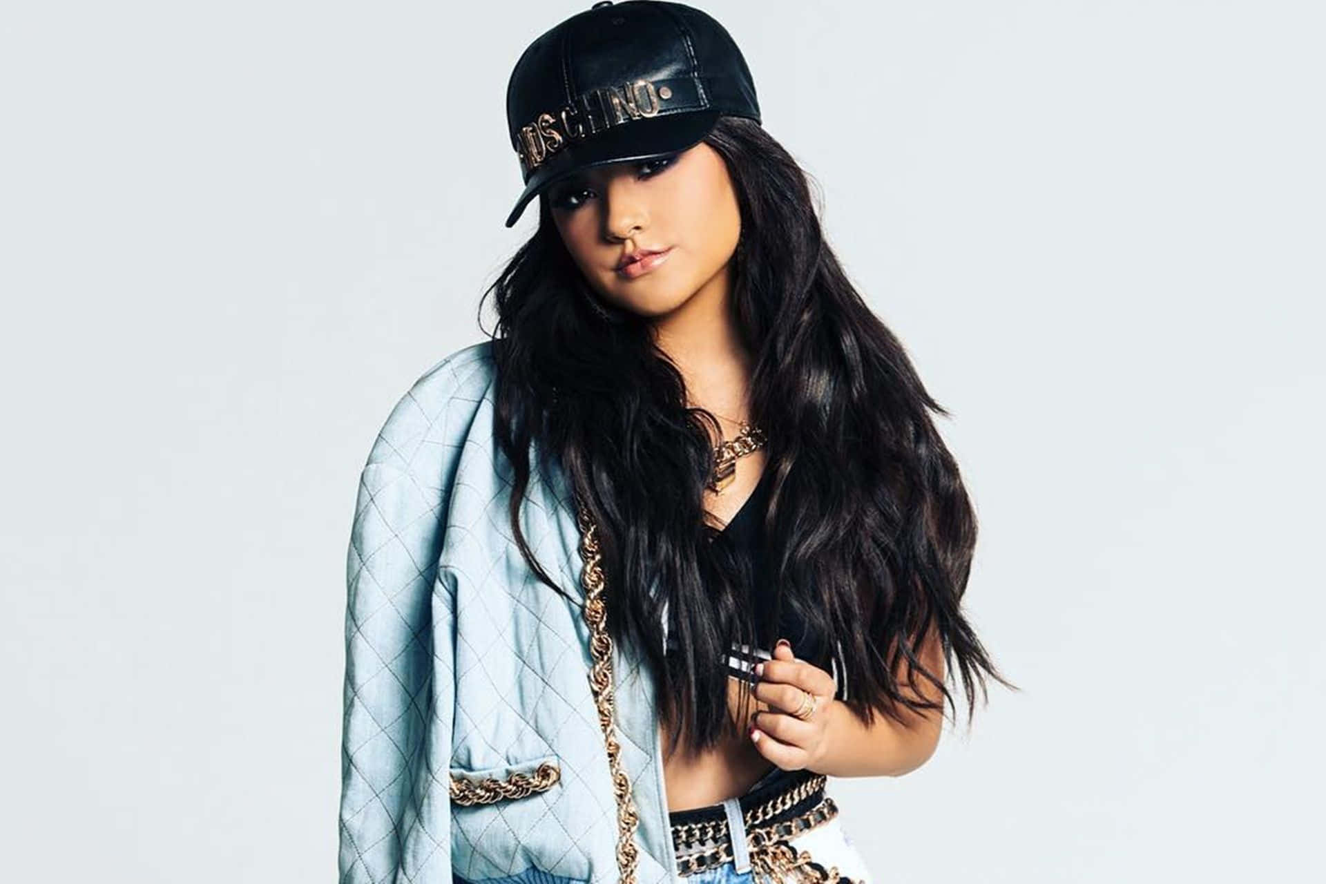 Becky G 桌布 壁紙 — 一位時尚的年輕女性，長長的黑髮，戴著黑色帽子，穿著淺藍色絎縫外套，背景為純白色。