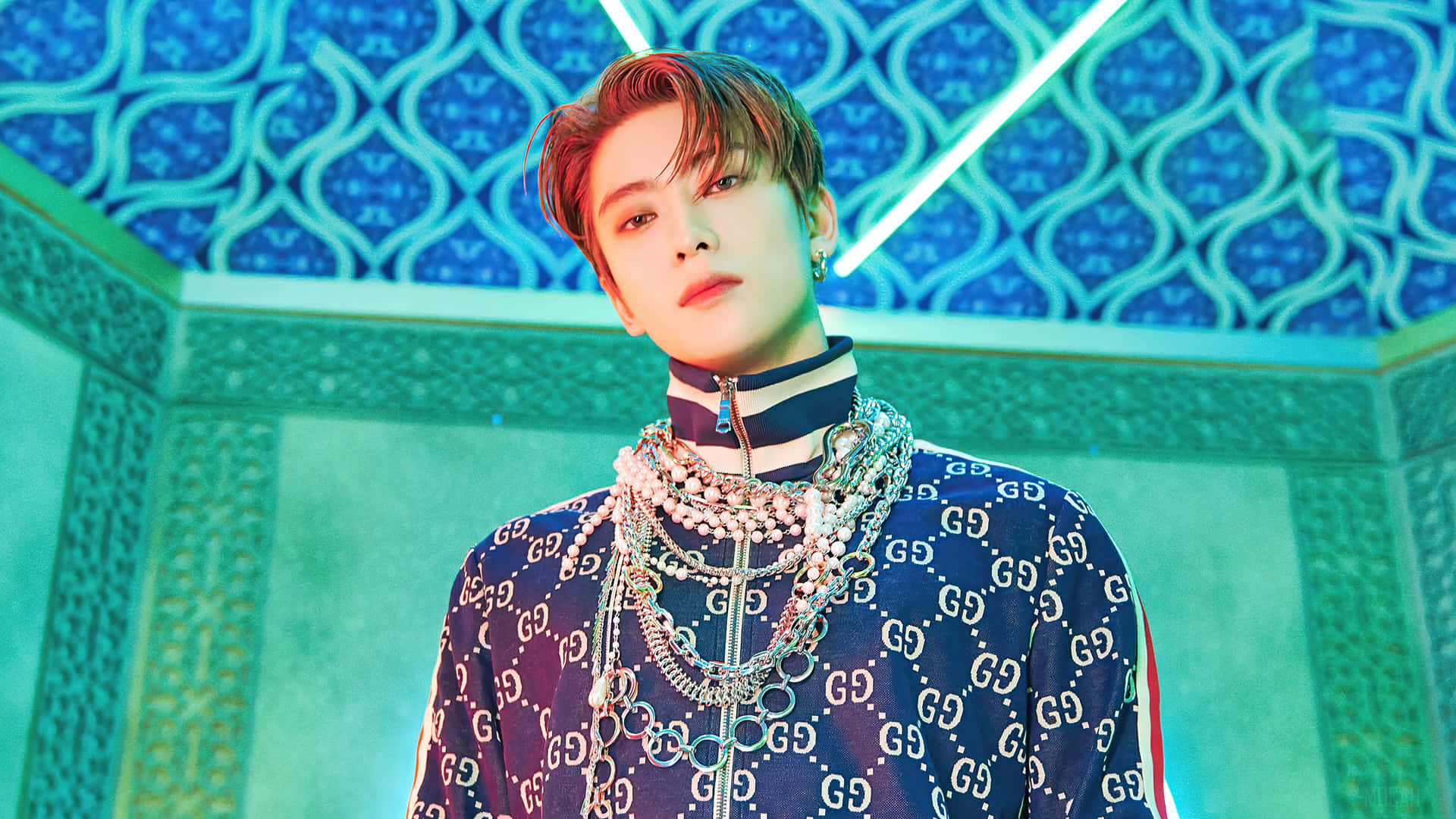 Gucci 桌布 壁紙 — 一位時尚的年輕男士身穿Gucci圖案服裝，佩戴多層項鍊，自信地站在藍色幾何圖案背景前。