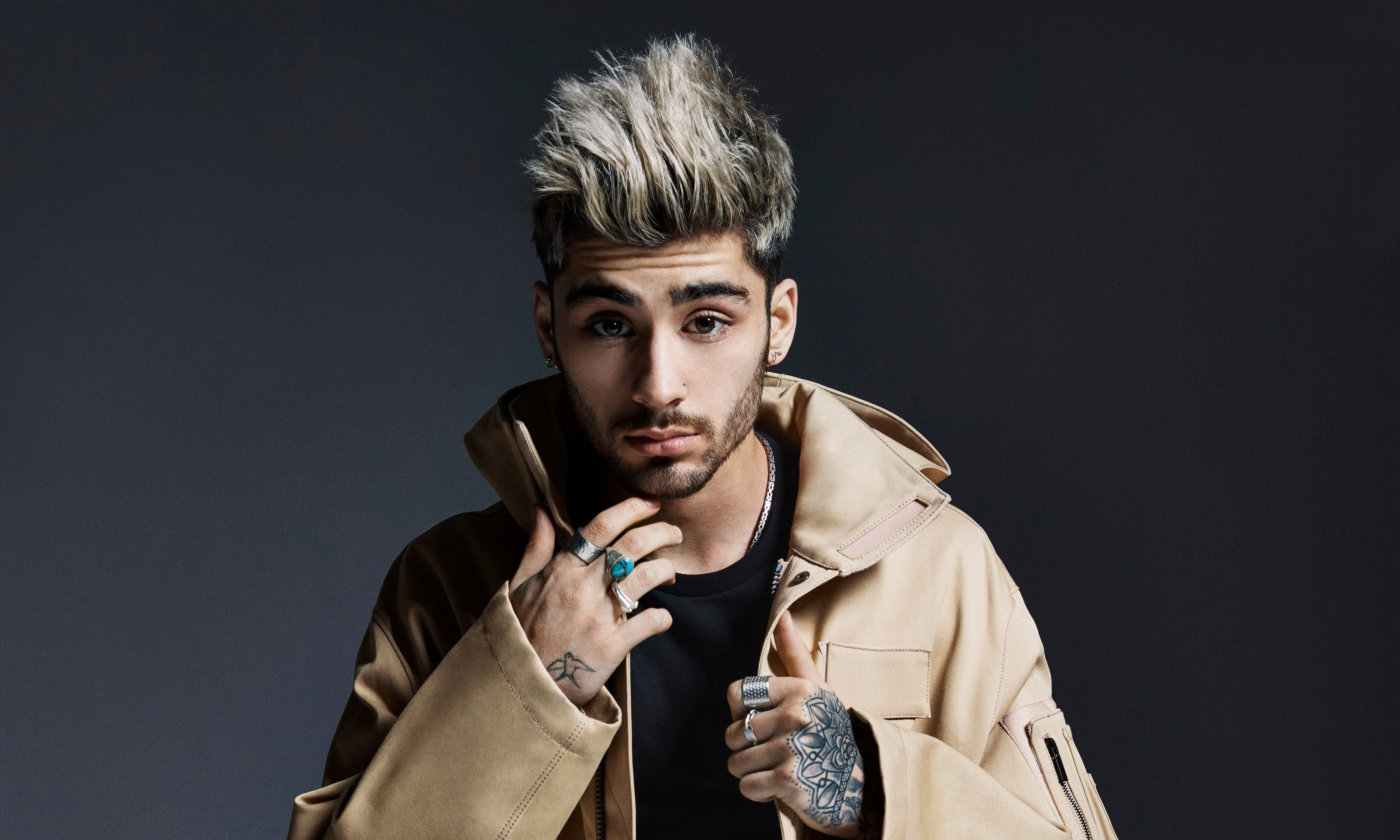 zayn 桌布 壁紙 — 一位時尚的年輕男子，有著尖刺金髮和刺青，穿著米色外套，在深色攝影棚背景前自信地擺姿勢。