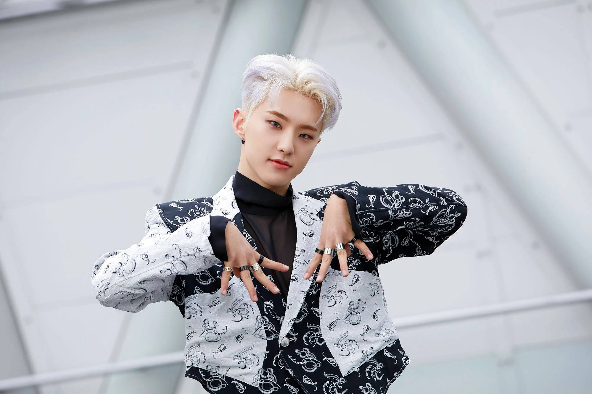 Hoshi 桌布 壁紙 — 一位時尚的年輕男子，留著鉑金色頭髮，穿著黑白圖案西裝外套，在現代白色建築背景下自信地擺出姿勢。