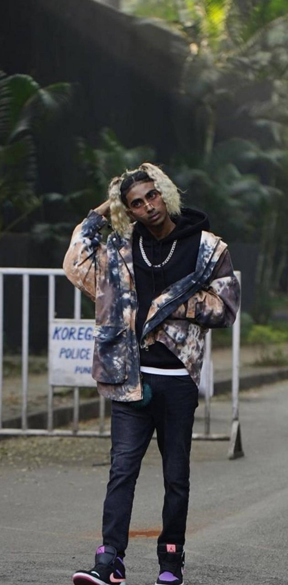 Celebrity 桌布 壁紙 — 一位時尚的年輕男子，金色辮子頭和眼鏡，在綠意盎然的城市小巷中，靠近警察路障，自信地穿著紮染外套擺姿勢。