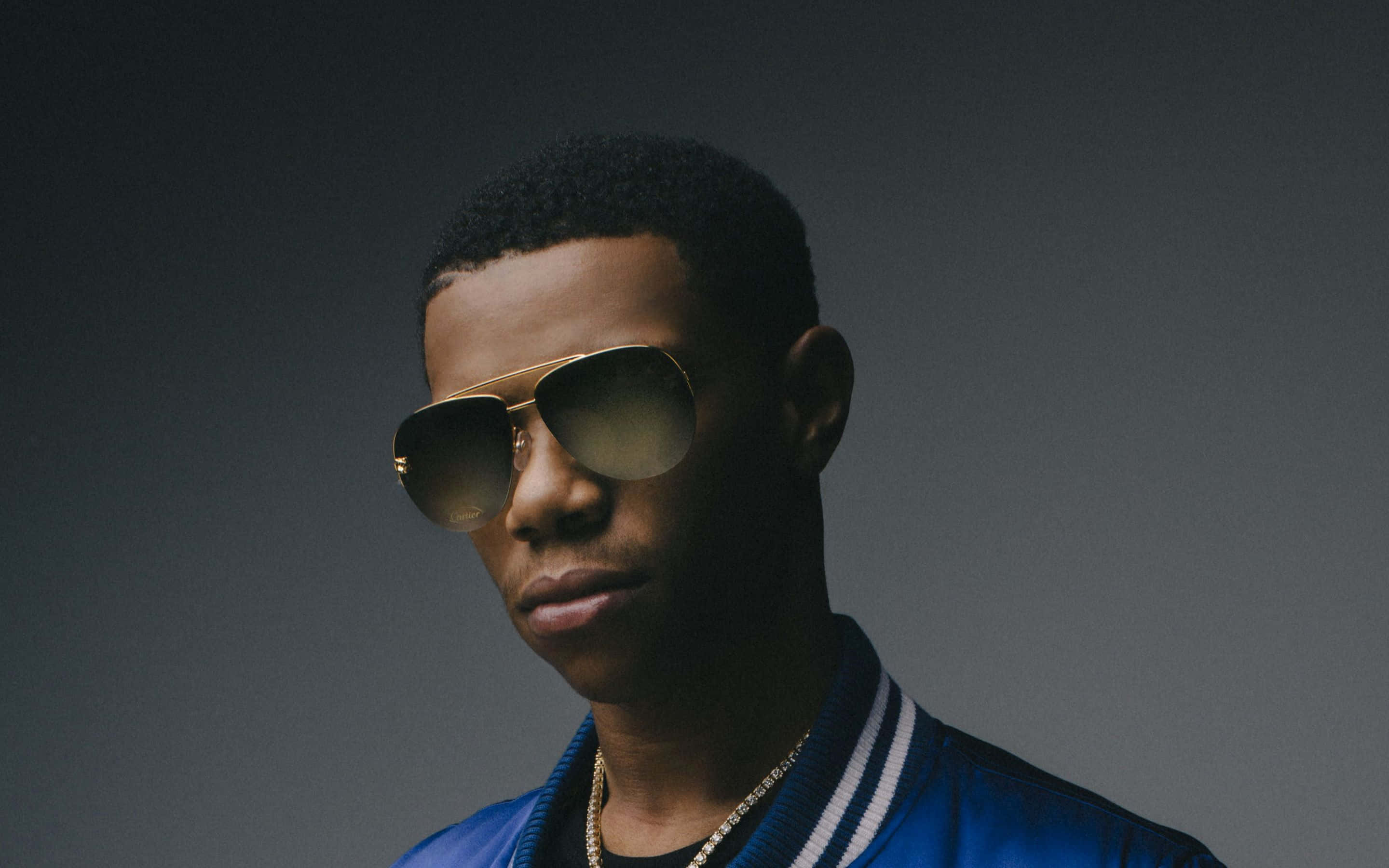 Rappers 桌布 壁紙 — 一位時尚的年輕男子戴著金色飛行員太陽眼鏡和藍色校隊夾克，在深灰色工作室背景前擺姿勢。