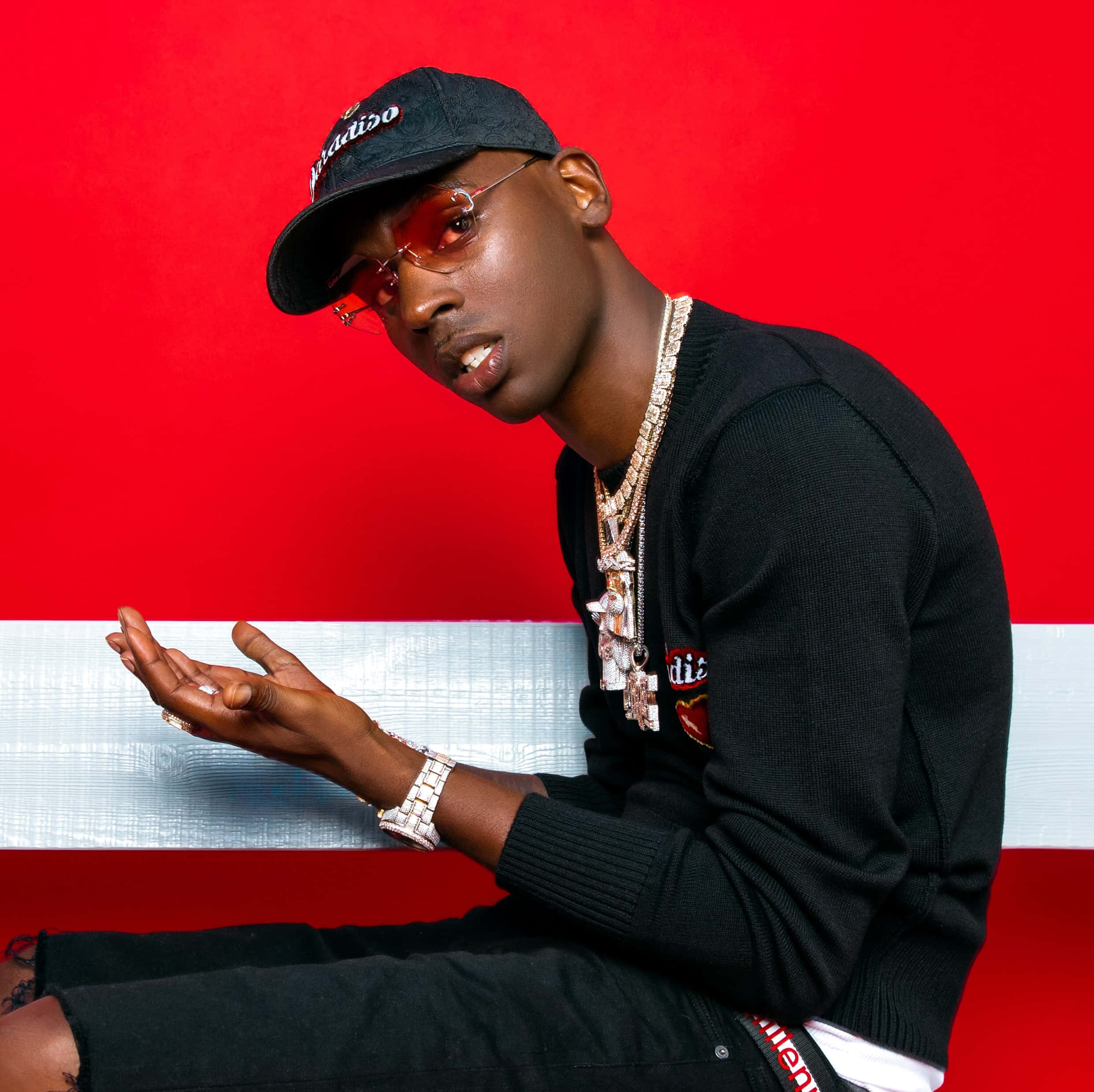 Young Dolph 桌布 壁紙 — 一位時尚的年輕男子戴著黑色鴨舌帽和紅色太陽眼鏡，在搶眼的紅白條紋背景前自信地擺姿勢。