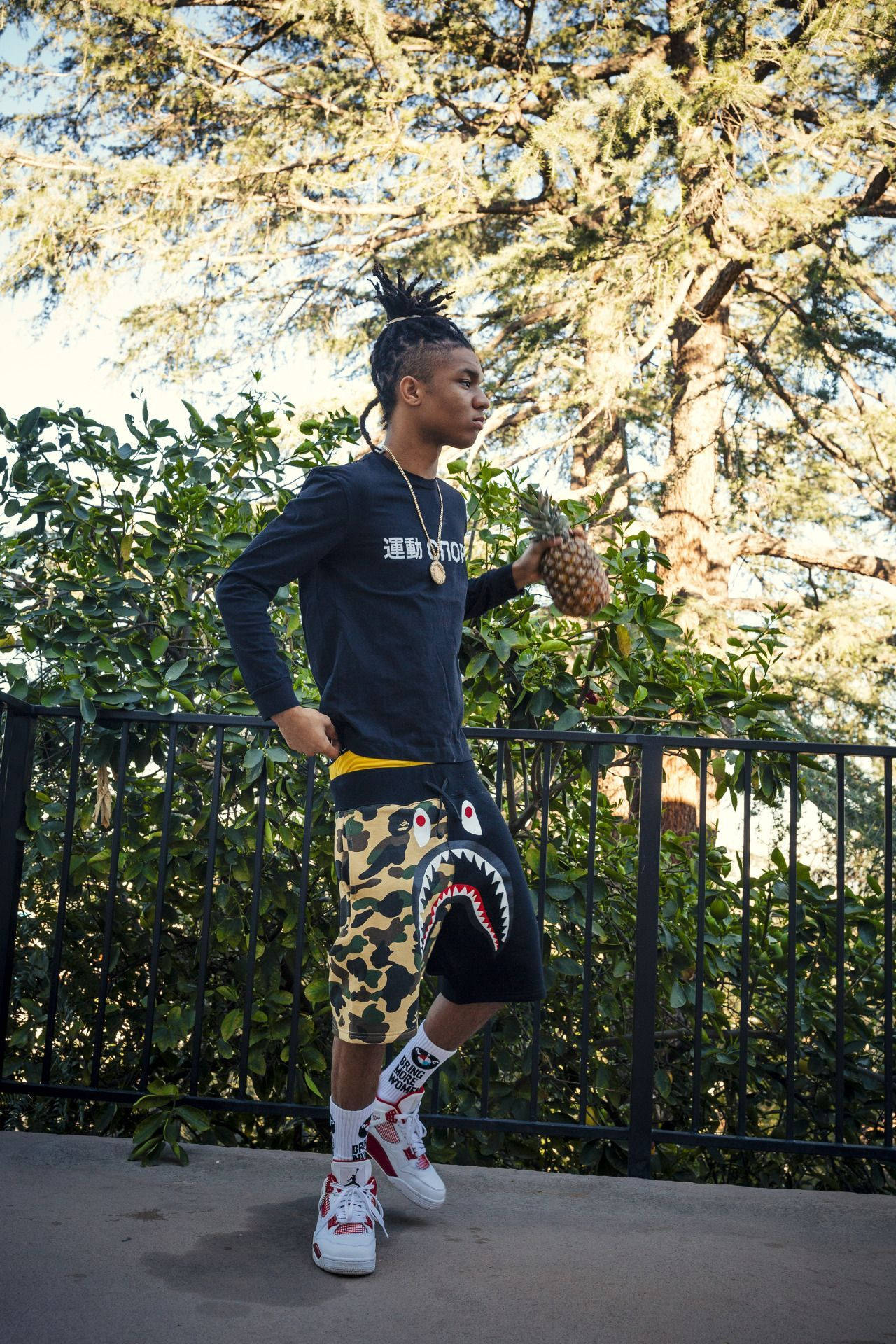 Swae Lee 桌布 壁紙 — 一位時尚的年輕男子梳著辮子，自信地站在陽台上，拿著一個鳳梨，穿著鯊魚印花短褲套裝。